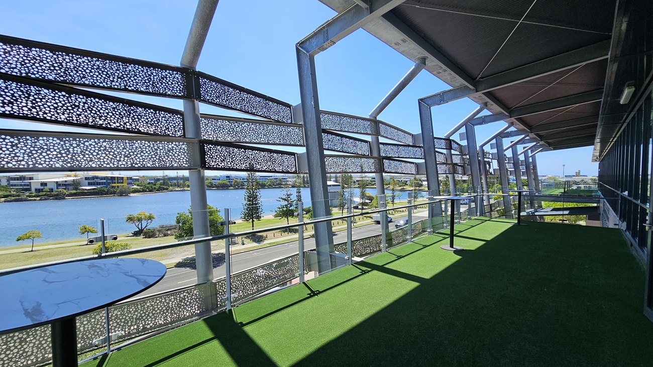 A balcony with artificial green grass flooring and a modern metal railing. The left side features decorative metal panels with circular cutouts. Two round tables are positioned along the railing. In the background, a river is visible, lined with trees and a road. Buildings are seen across the river. The ceiling has a metal framework with a light fixture.
