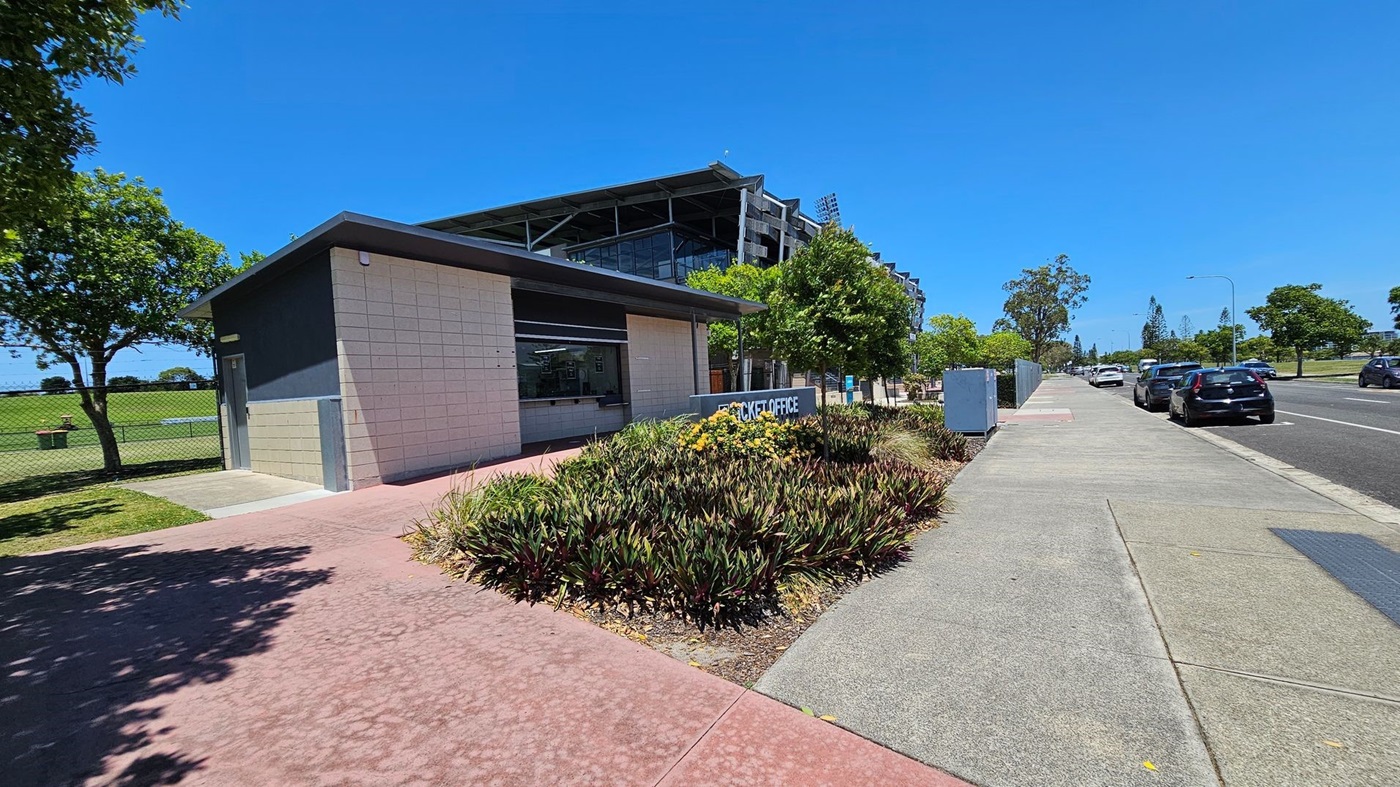 A small building with a flat roof is on the left, featuring a sign that reads "TICKET OFFICE" in front of a garden with yellow flowers and green plants. Behind it, a larger structure with a modern design is visible. A tree and a grassy area are to the left, enclosed by a fence. On the right, a road with parked cars runs alongside a pavement lined with trees and utility boxes. The sky is clear and blue.