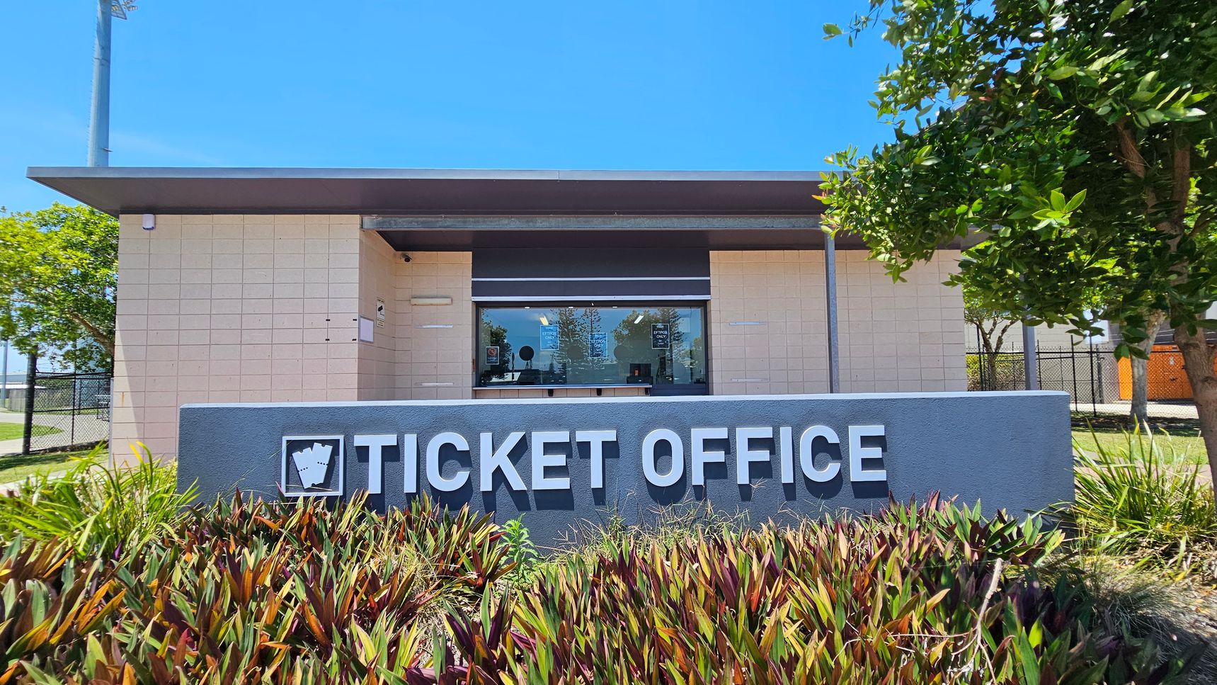 A ticket office building with a flat roof and beige brick walls. In the foreground, a grey sign with the words "TICKET OFFICE" is surrounded by green and reddish-brown plants. On the left, a tall light pole and a tree are visible. On the right, another tree provides shade. The window of the ticket office is in the centre, with some posters and notices visible inside.
