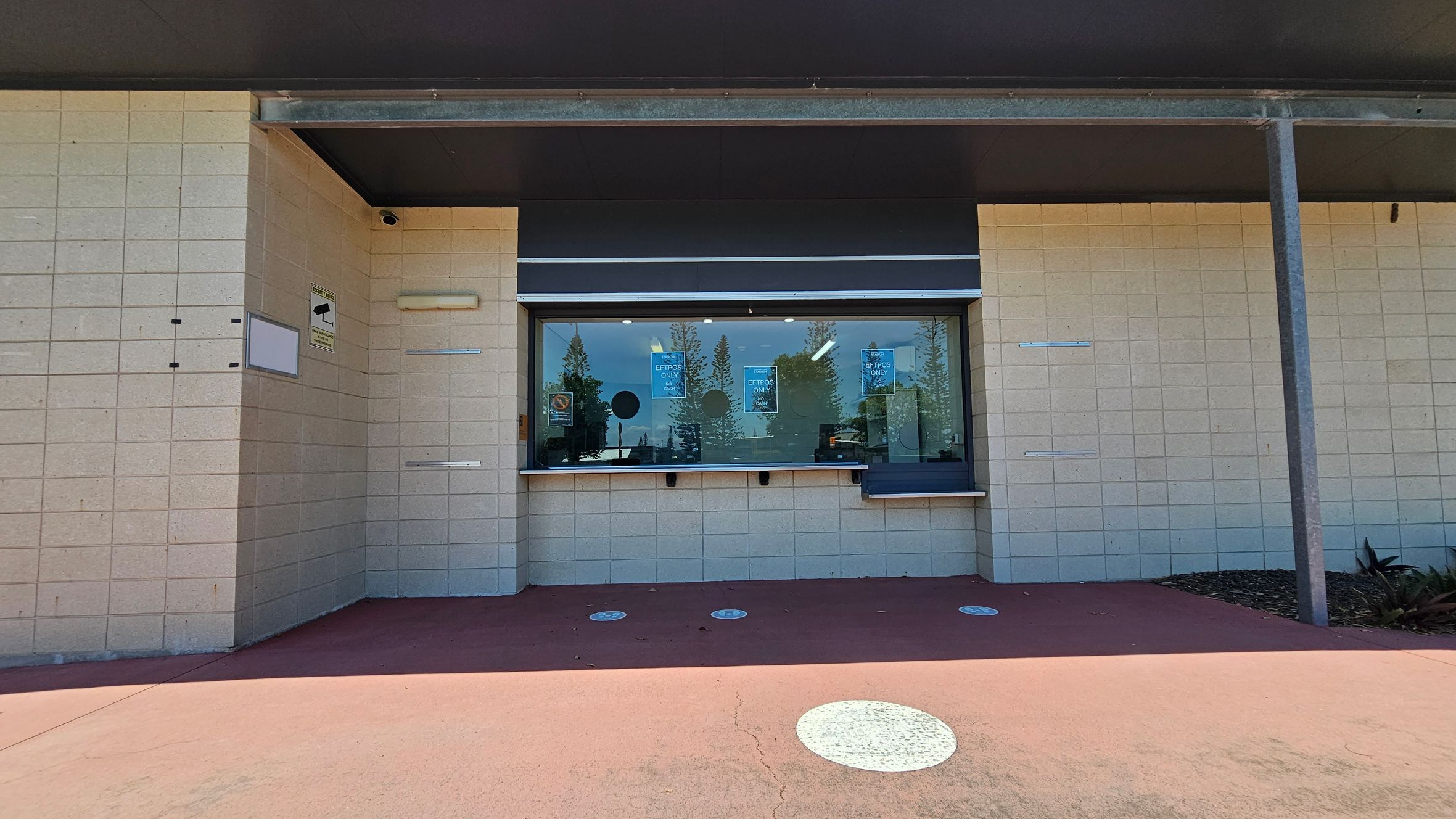 A building facade with a large glass window in the centre, featuring signs that read "EFTPOS ONLY." The wall is made of light-coloured bricks. On the left, there is a security camera sign and a small camera above. The ground is a reddish-brown concrete with circular white markings. The ceiling is dark and supported by a metal beam on the right. Some plants are visible at the bottom right corner.