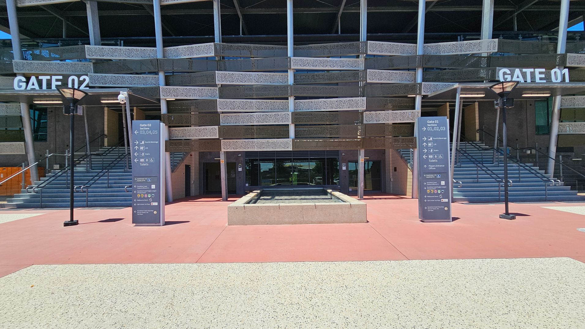 The image shows the entrance to Sunshine Coast Stadium. In the centre, there are steps leading up to Gate 01, with a sign above. To the left is Gate 02. The foreground features a metal fence with an open gate, and a sign detailing the Kawana Sports Precinct conditions of entry. The ground is a mix of red and light-coloured paving. The stadium's facade has a modern design with horizontal slats and large windows.