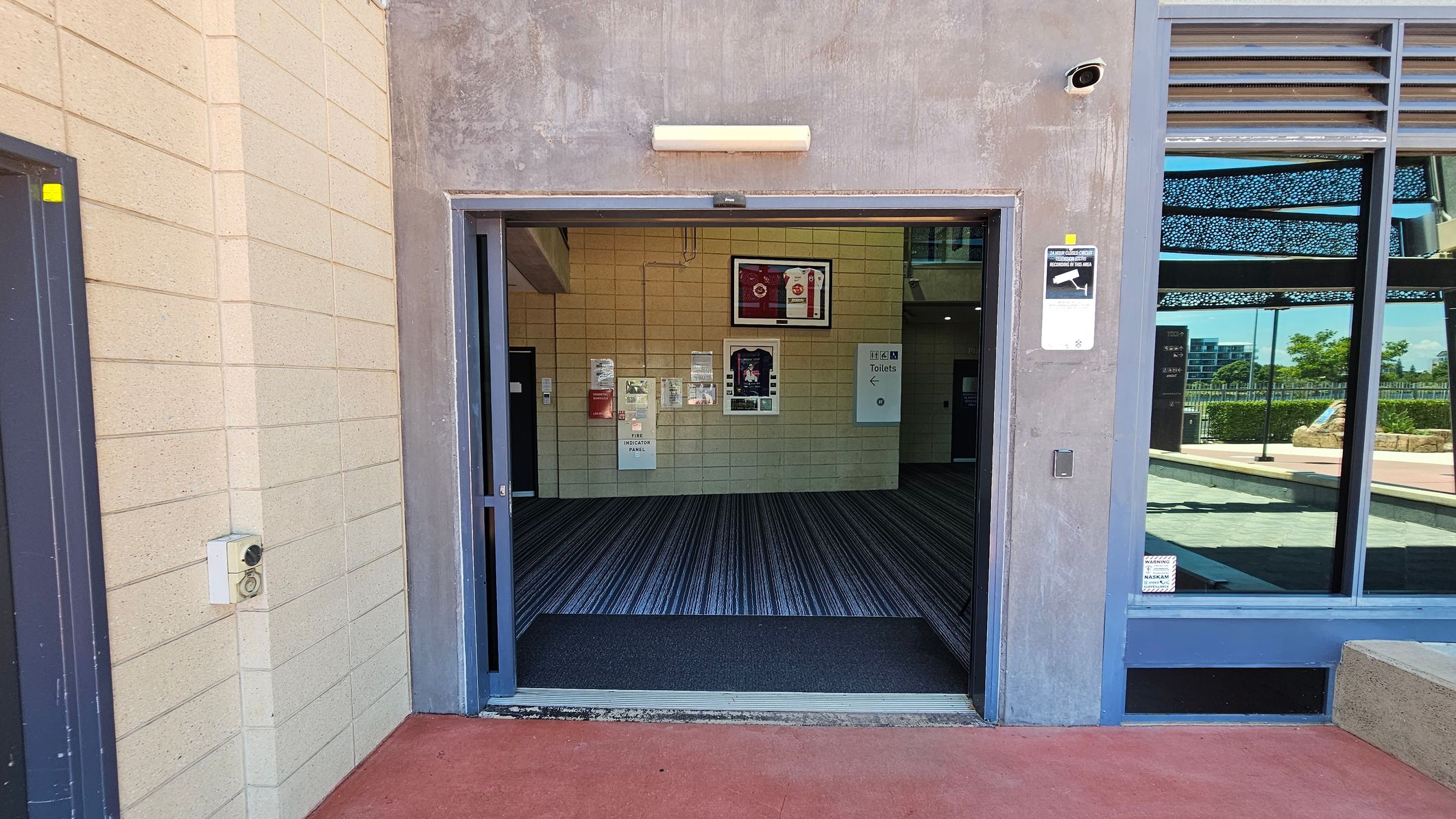 A modern building entrance with two gates labelled "Gate 01" and "Gate 02" on the upper level. In the centre, a rectangular water feature is surrounded by a concrete border. Two tall signposts with directional information stand on either side of the water feature. Wide staircases lead up to the gates, flanked by metal railings. The ground is a mix of red and light grey paving. The building facade features perforated metal panels and large glass doors.
