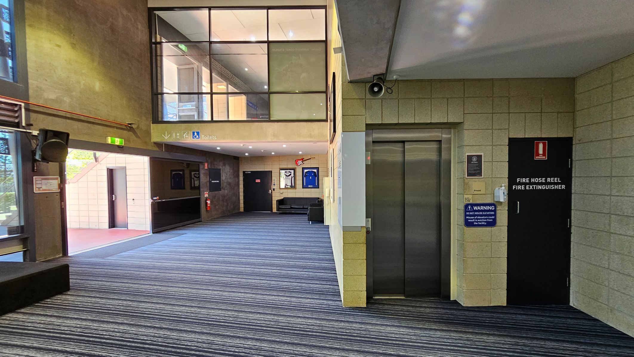 A spacious indoor area with a striped carpeted floor. On the left, there's a sign indicating the toilets, and framed sports jerseys are displayed on the walls. The right side features large glass windows and a black platform. The upper level has a glass railing and more framed items. The ceiling is dotted with recessed lights, and a central pillar supports the structure.