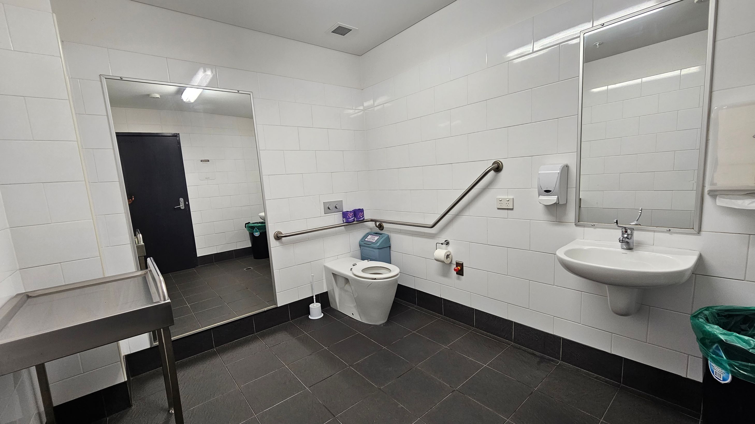 A public bathroom with white tiled walls and a dark tiled floor. On the right, there's a wall-mounted sink with a mirror above it, a soap dispenser, and a paper towel holder. In the centre, a toilet with a support rail and a sanitary bin nearby. On the left, a large mirror reflects the opposite wall and a black door. A stainless steel table is partially visible. A green-lined bin is on the floor near the sink. The ceiling is plain with a fluorescent light fixture.