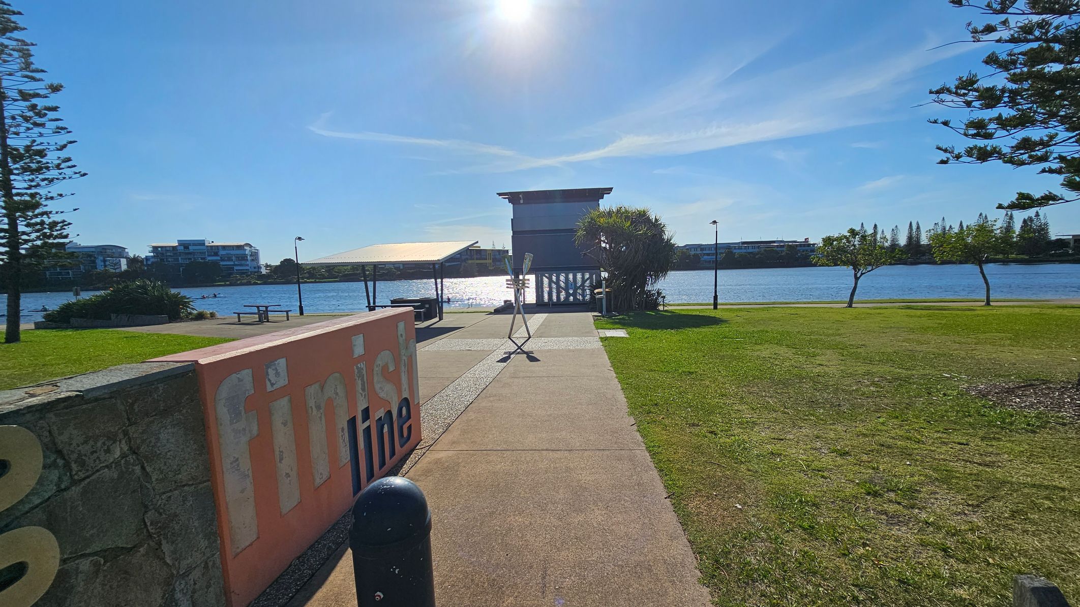 A riverside park with a concrete path leading to a structure by the water. On the left, a large sign reads "finish line" next to a stone wall. The grass is neatly trimmed, with a few trees scattered around. In the background, the river reflects the sunlight, and there are buildings on the opposite bank. A covered seating area is visible near the water, and a tall pole stands in the centre of the path.