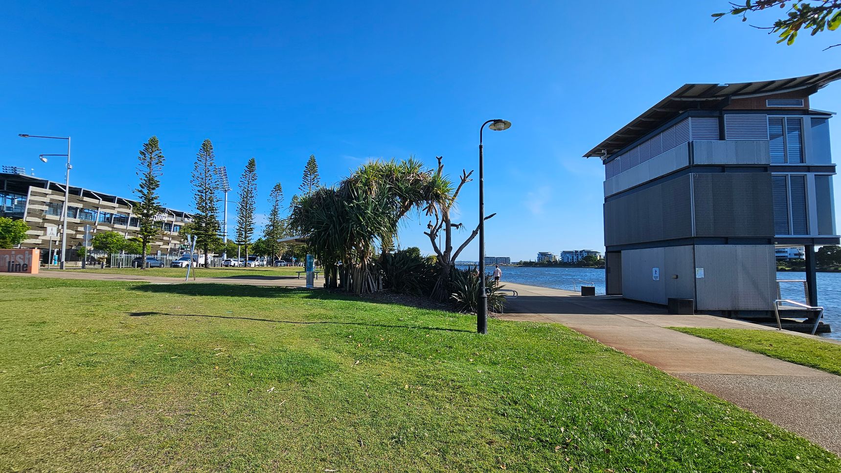 A grassy area with a path runs along the left side, bordered by a row of tall trees and a modern building. In the centre, a cluster of large, leafy plants grows near a lamppost. On the right, a two-storey building with large windows stands beside a body of water. A person sits on a bench by the water, and distant buildings are visible across the water. The sky is clear and blue.