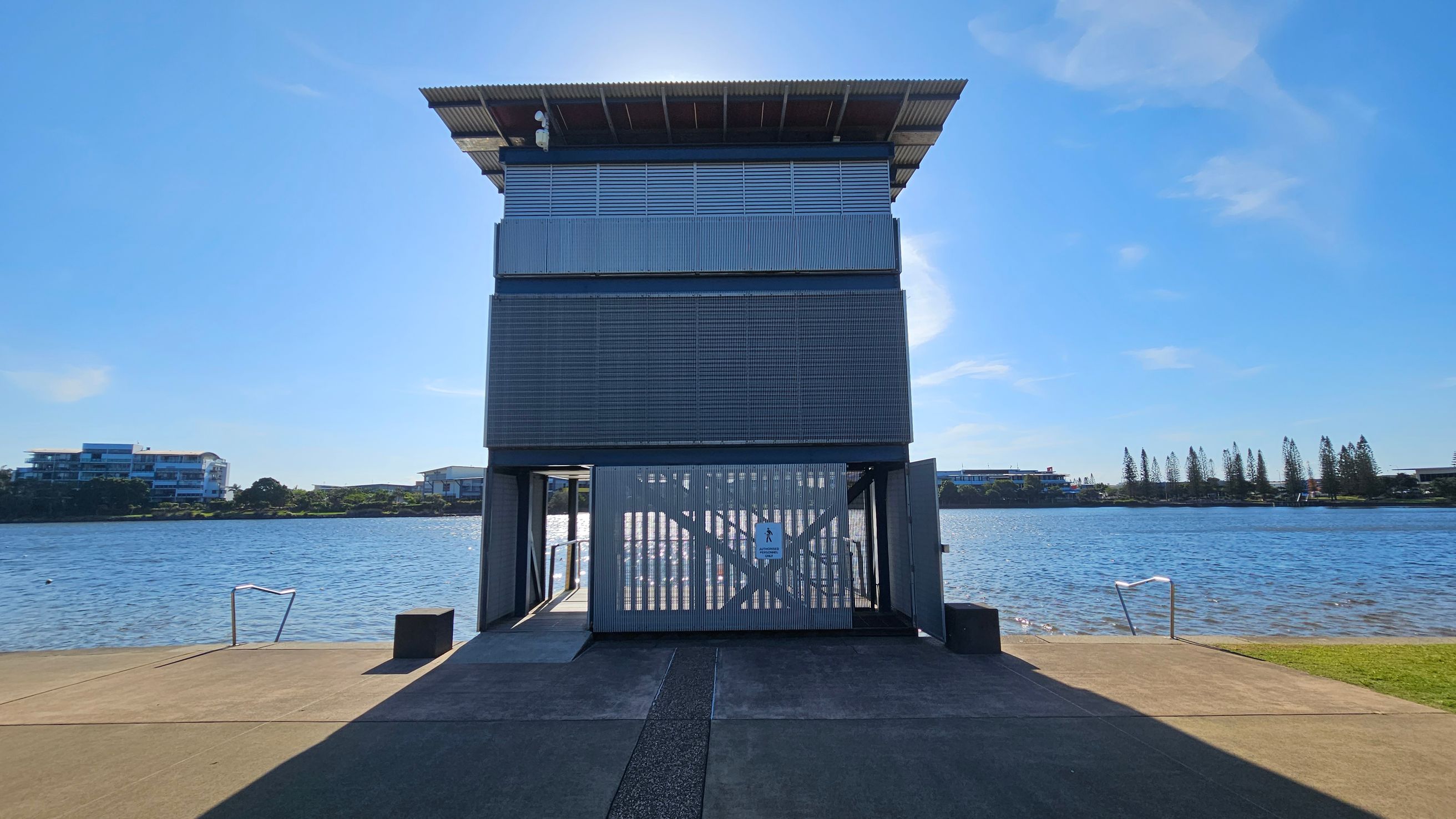A modern, rectangular structure with metal cladding stands on a concrete platform by a body of water. The building has a corrugated metal roof and a gated entrance with a pedestrian sign. In the background, across the water, are low-rise buildings and a line of trees. The foreground features a concrete path leading to the structure, with metal railings descending into the water on either side.