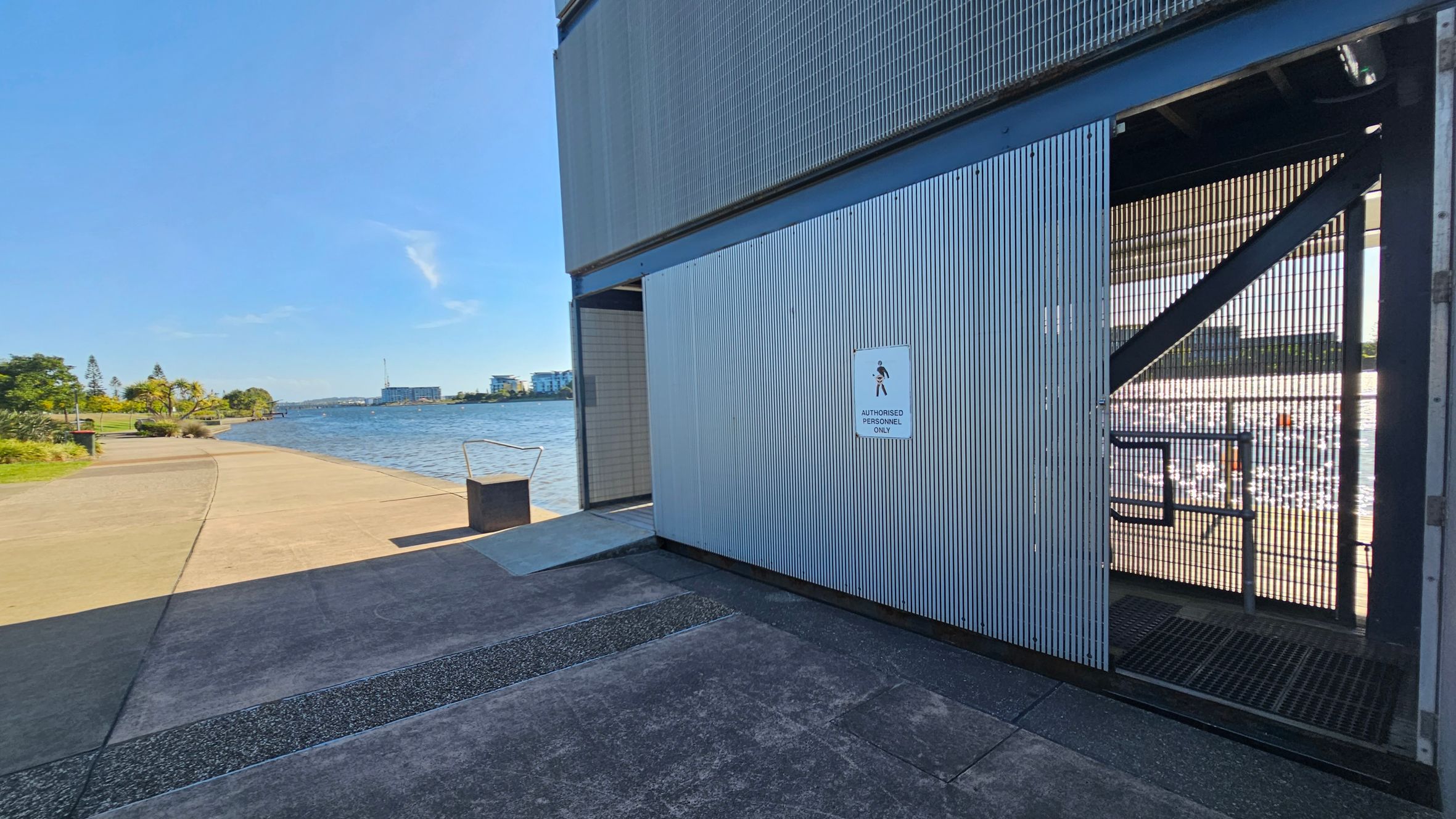 A riverside walkway with a concrete path on the left, bordered by greenery and trees. The river is visible, with buildings in the distance. On the right, a large metal structure with a sign reading "Authorised Personnel Only" is partially open, revealing a grated floor inside. A metal bin is positioned near the edge of the path. The sky is clear and blue.