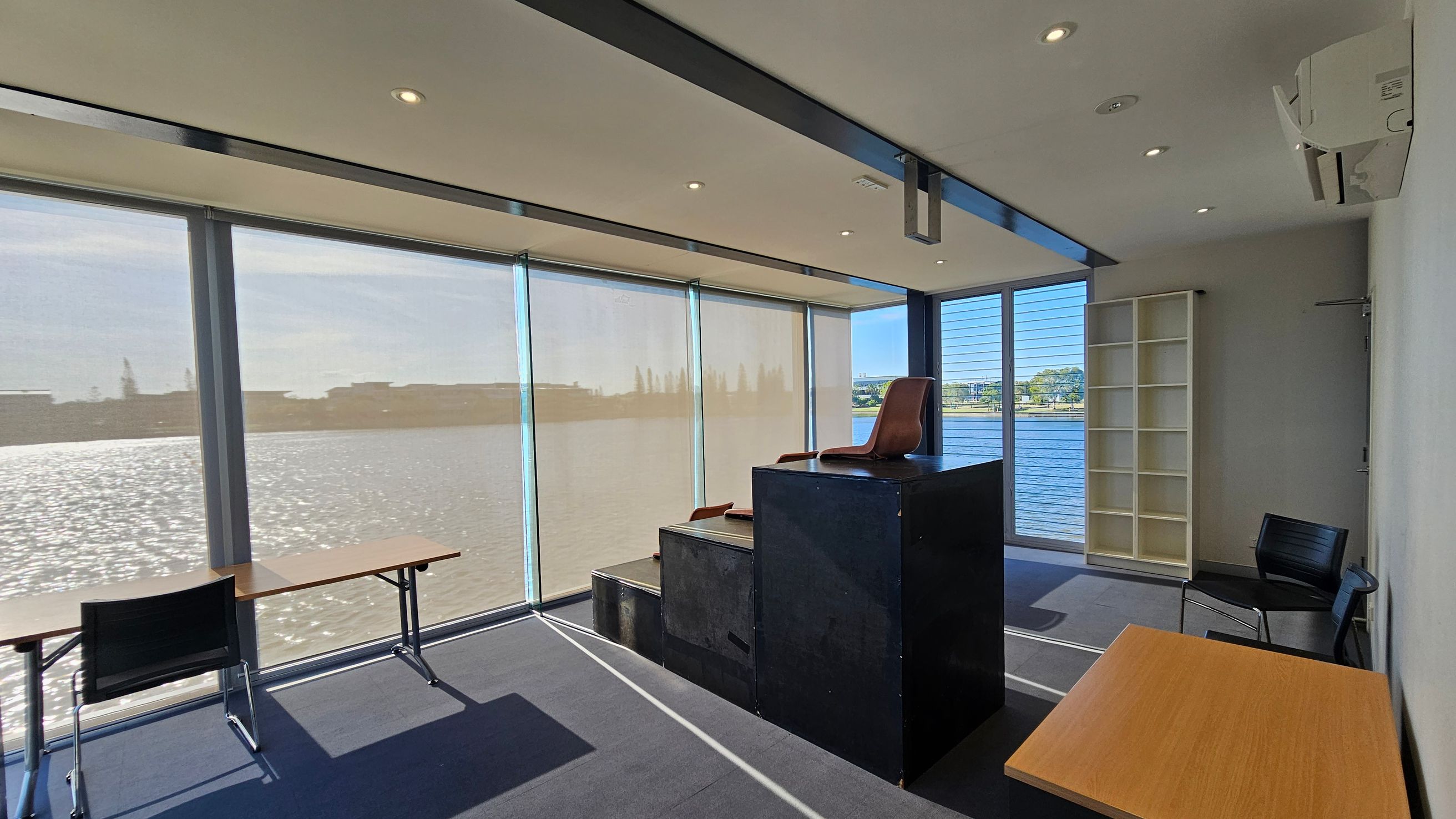 A room with large windows overlooking a body of water. On the left, there's a long table with a black chair. In the centre, a raised platform with a brown chair on top. On the right, a white shelving unit and a black chair. The floor is carpeted in grey, and the ceiling has recessed lighting and an air conditioning unit.