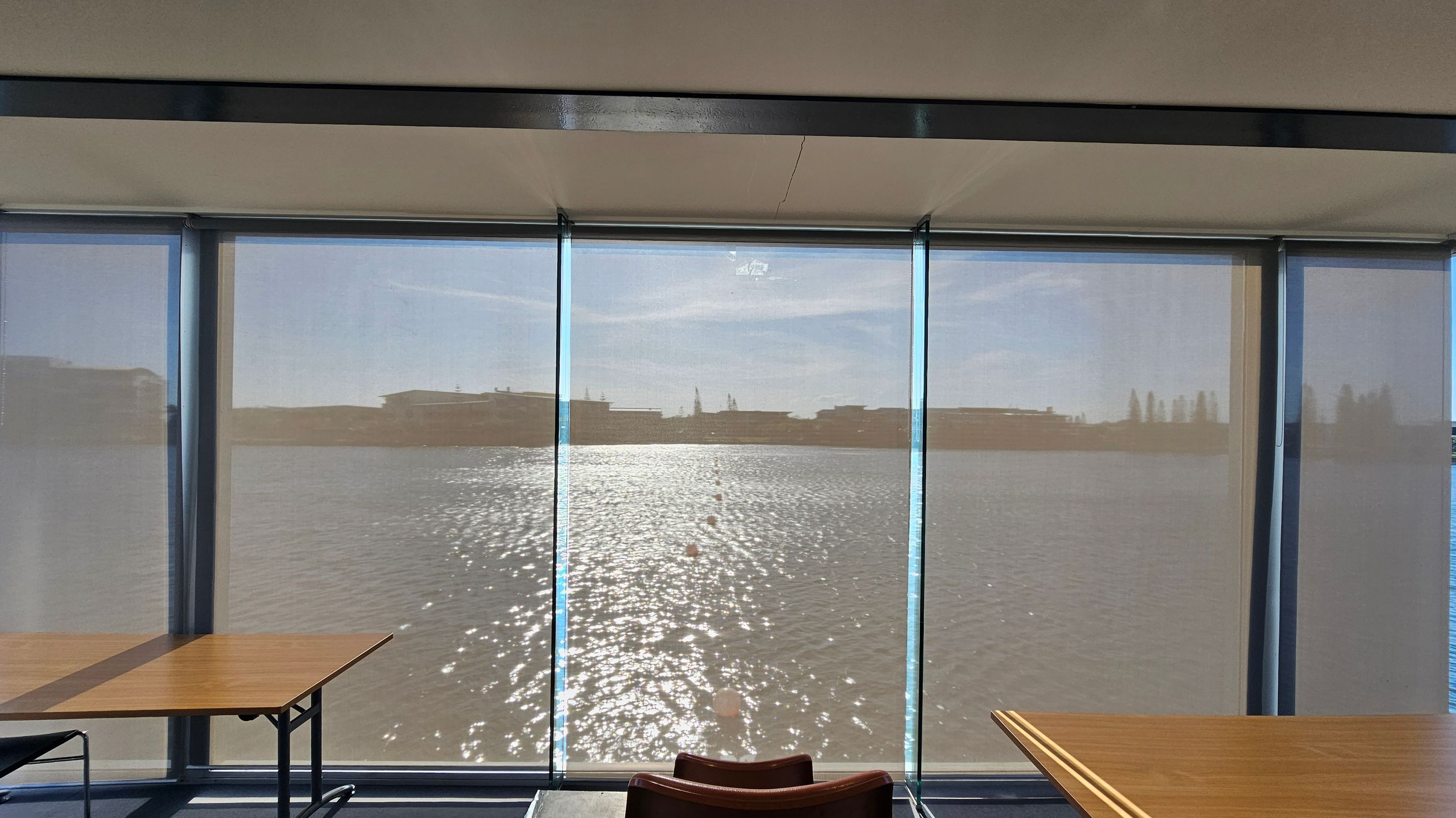 Large windows with sheer blinds cover the entire wall, offering a view of a body of water with sunlight reflecting off its surface. In the foreground, there are two wooden tables with metal legs, one on the left and one on the right. A chair is partially visible between the tables. The ceiling is plain and light-coloured, with a dark horizontal beam running across the top of the windows.