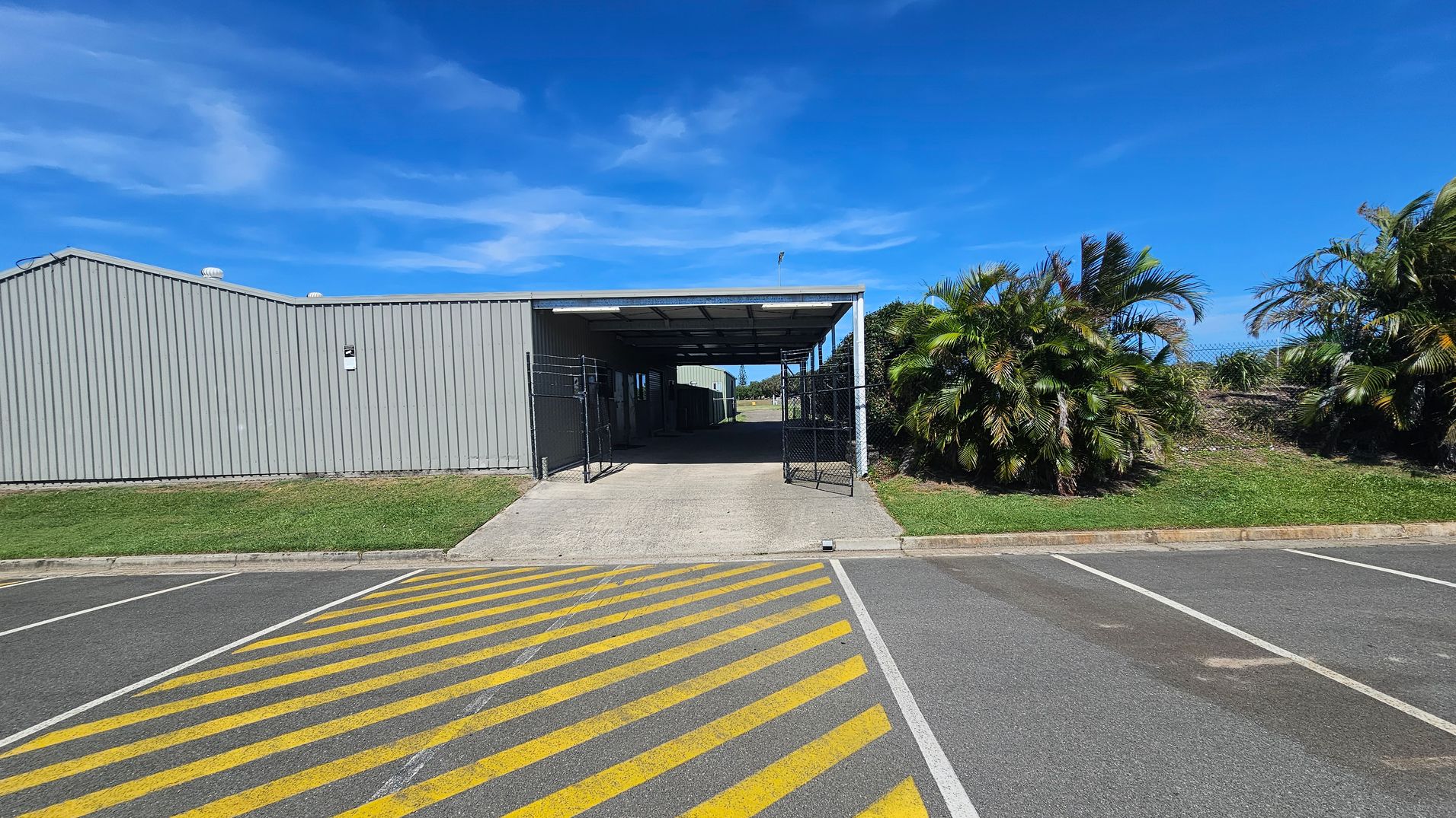 A grey corrugated metal building with a covered entrance is in the centre. The entrance has open black metal gates. To the right, there are lush green palm trees and grass. The foreground features a car park with yellow diagonal lines painted on the asphalt. The sky is clear and blue.