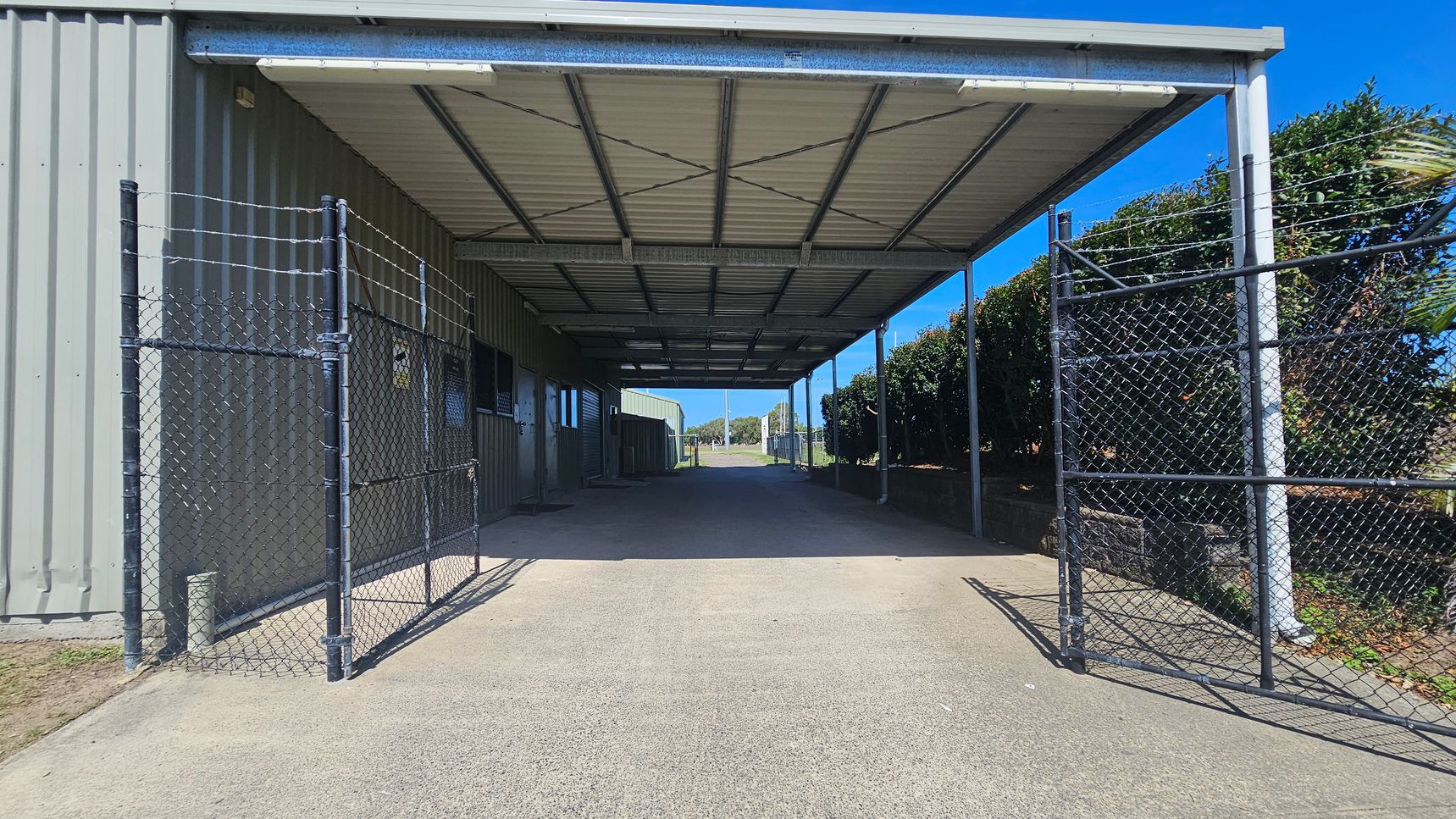 A covered walkway with a metal roof and chain-link fencing on both sides. The left side has a corrugated metal wall with doors and windows, while the right side features a row of bushes. The ground is paved, and the ceiling has metal beams. The walkway leads to an open area in the distance.