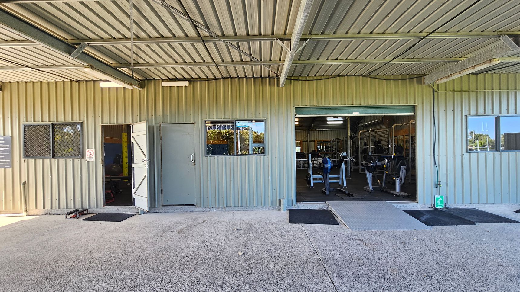 A corrugated metal building with an open entrance on the right, revealing gym equipment inside. There are two small windows with metal grilles, one on each side of the entrance. A closed grey door is next to the left window. The floor is concrete with black mats near the entrances. The ceiling is also corrugated metal with visible beams. A green emergency exit sign is on the wall near the open entrance.