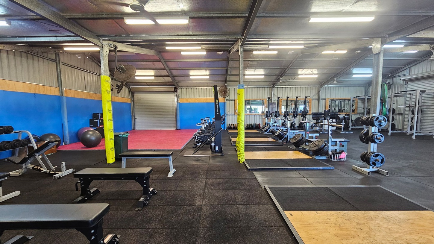 A spacious gym interior with a variety of equipment. On the left, there are benches and dumbbells on a rack, with exercise balls nearby. The floor is covered with black rubber mats. In the centre, rowing machines are lined up, and a red mat area is visible in the background. On the right, there are weightlifting platforms and racks with weight plates. The walls are blue and grey, and the ceiling has fluorescent lights.