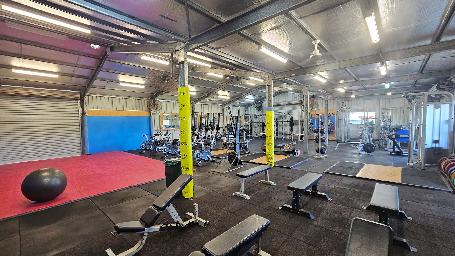 A spacious gym with a high metal ceiling and fluorescent lights. On the left, there's a red mat with a large black exercise ball. In the centre, several exercise bikes are lined up against a blue and yellow wall. The right side features weightlifting equipment, benches, and racks. The floor is covered with black rubber mats, and yellow padding wraps around metal support beams. The walls are made of corrugated metal, and there are large fans mounted on them.