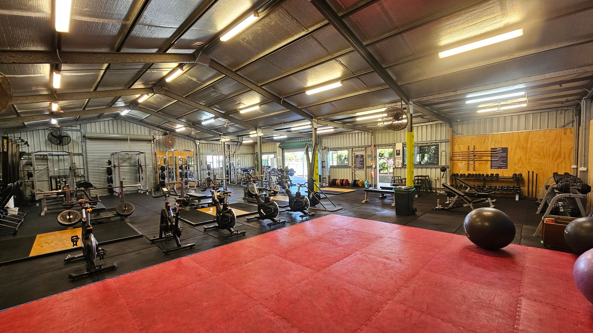 A spacious gym interior with a high metal ceiling and fluorescent lighting. On the left, there are weightlifting racks and benches. In the centre, several stationary bikes are lined up on a black mat. The right side features a wall with dumbbells and exercise equipment. The floor has a large red mat area in the foreground, and exercise balls are scattered around. Windows and a partially open roller door are visible in the background, allowing natural light in.