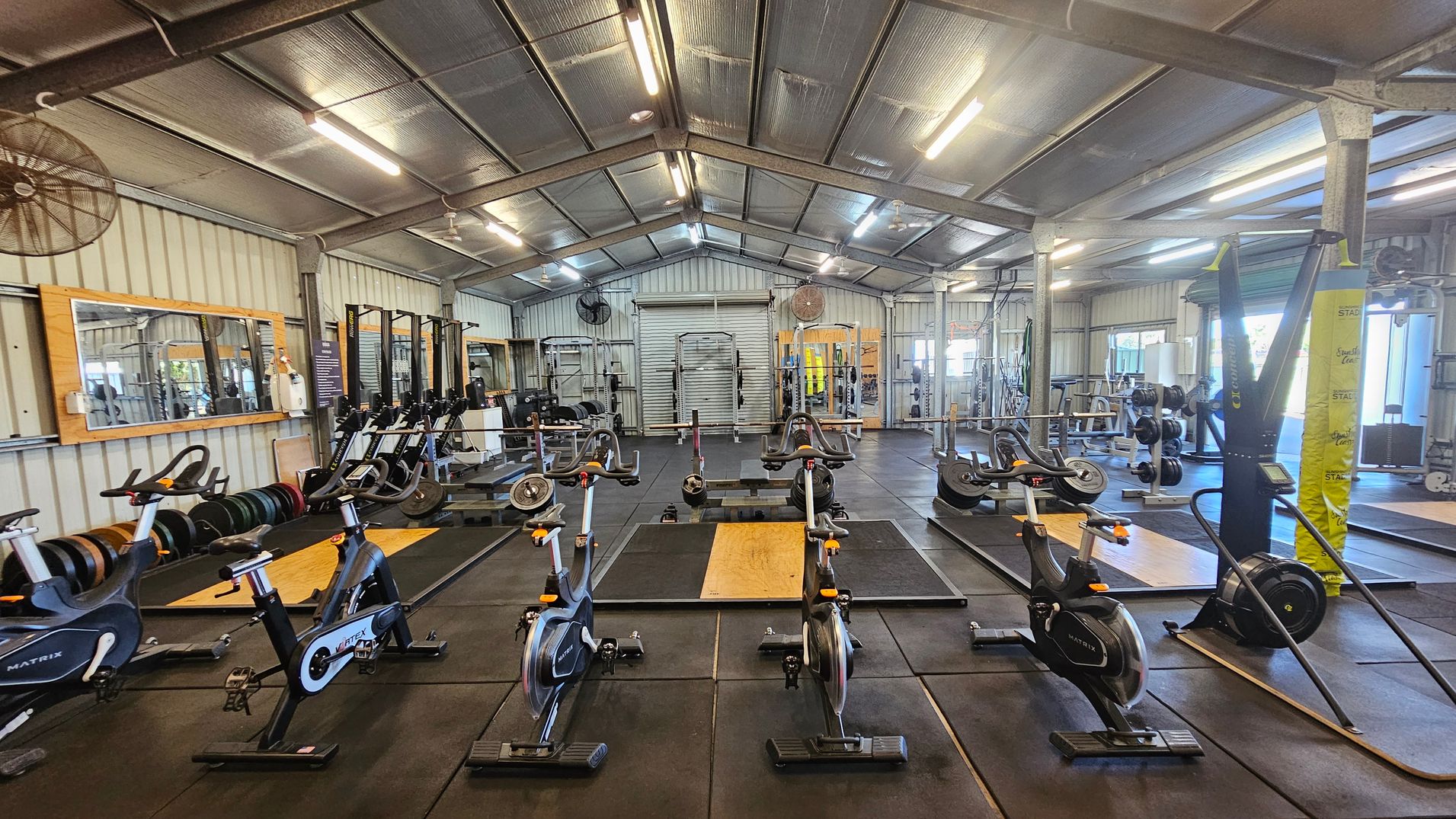 A spacious gym with a high metal ceiling and rubber flooring. In the foreground, several stationary bikes are lined up. In the middle, there are weightlifting platforms with barbells and weights. The background features a variety of gym equipment, including squat racks and benches. Large mirrors are mounted on the left wall, and there are industrial fans attached to the walls. The gym is well-lit with fluorescent lights.