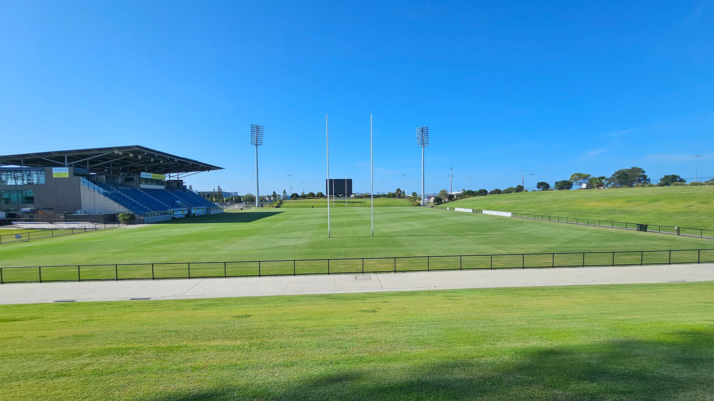 A rugby field with green grass and goalposts in the centre. On the left, there is a covered grandstand with blue seating. In the background, tall floodlights and a scoreboard are visible. The field is surrounded by a low fence, and there is a paved walkway in the foreground. Trees and open grassy areas are in the distance.