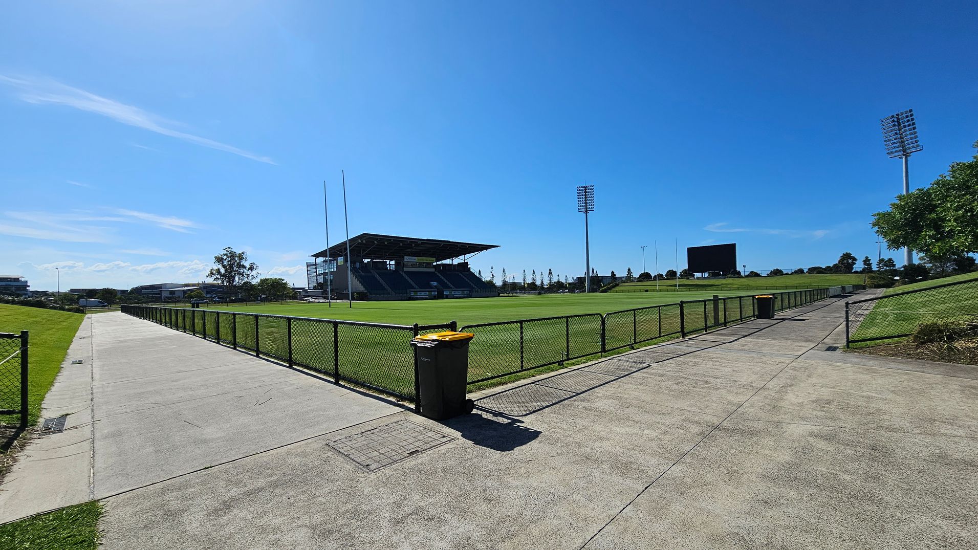 A sports field with a large, covered grandstand on the right. The field is surrounded by a black chain-link fence. A concrete path runs alongside the fence, with a yellow-lidded bin in the foreground. Tall floodlights are visible on the right, and a large scoreboard is in the distance. The grass is well-maintained, and there are trees and buildings in the background. The sky is clear and blue.