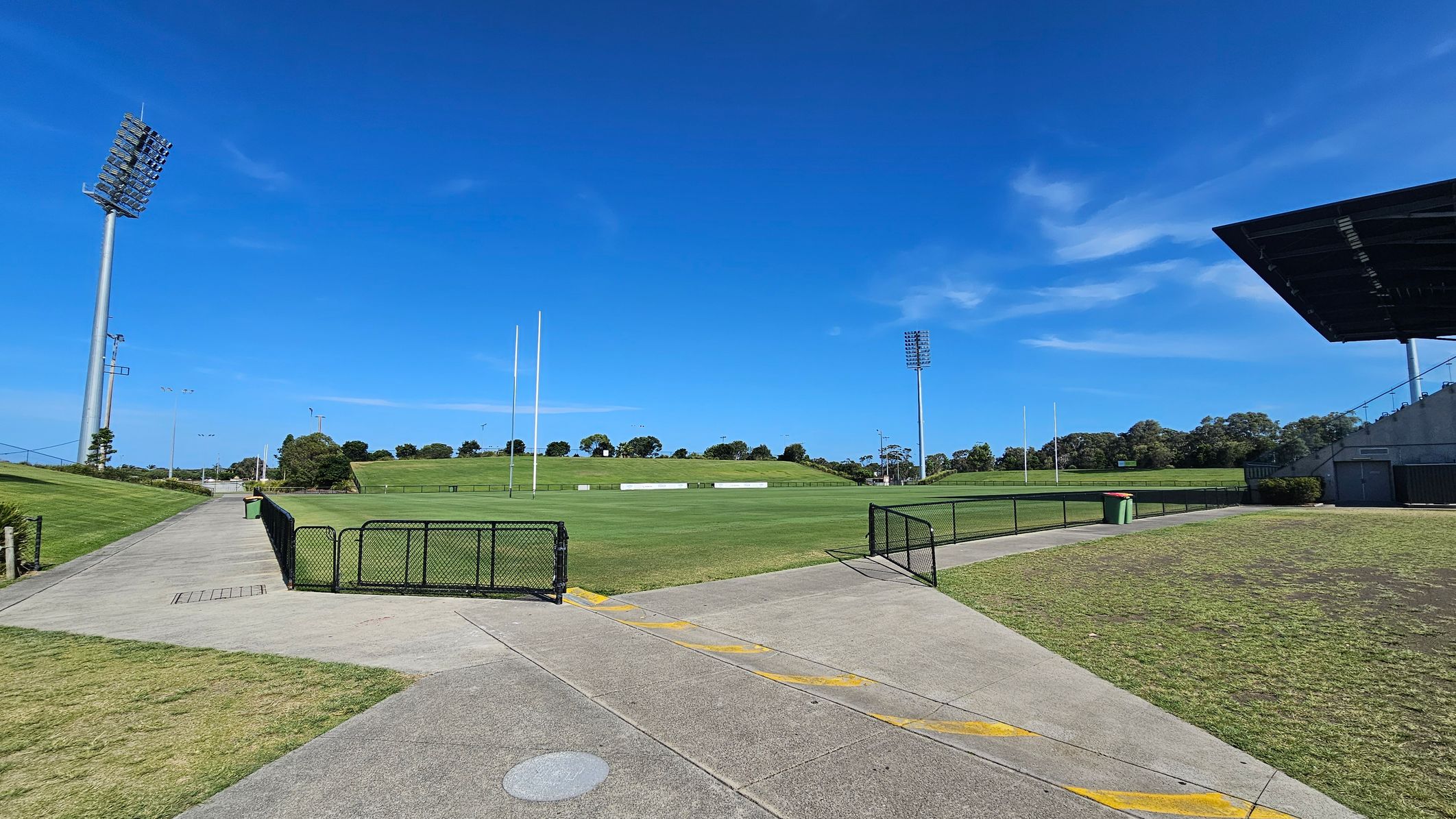 A sports field with green grass and rugby posts in the centre. On the left, a tall floodlight stands beside a paved walkway. On the right, a covered seating area with a slanted roof is visible. The foreground has a mix of grass and concrete paths, with a green bin near the seating area. Trees line the background, and another floodlight is visible on the right side.