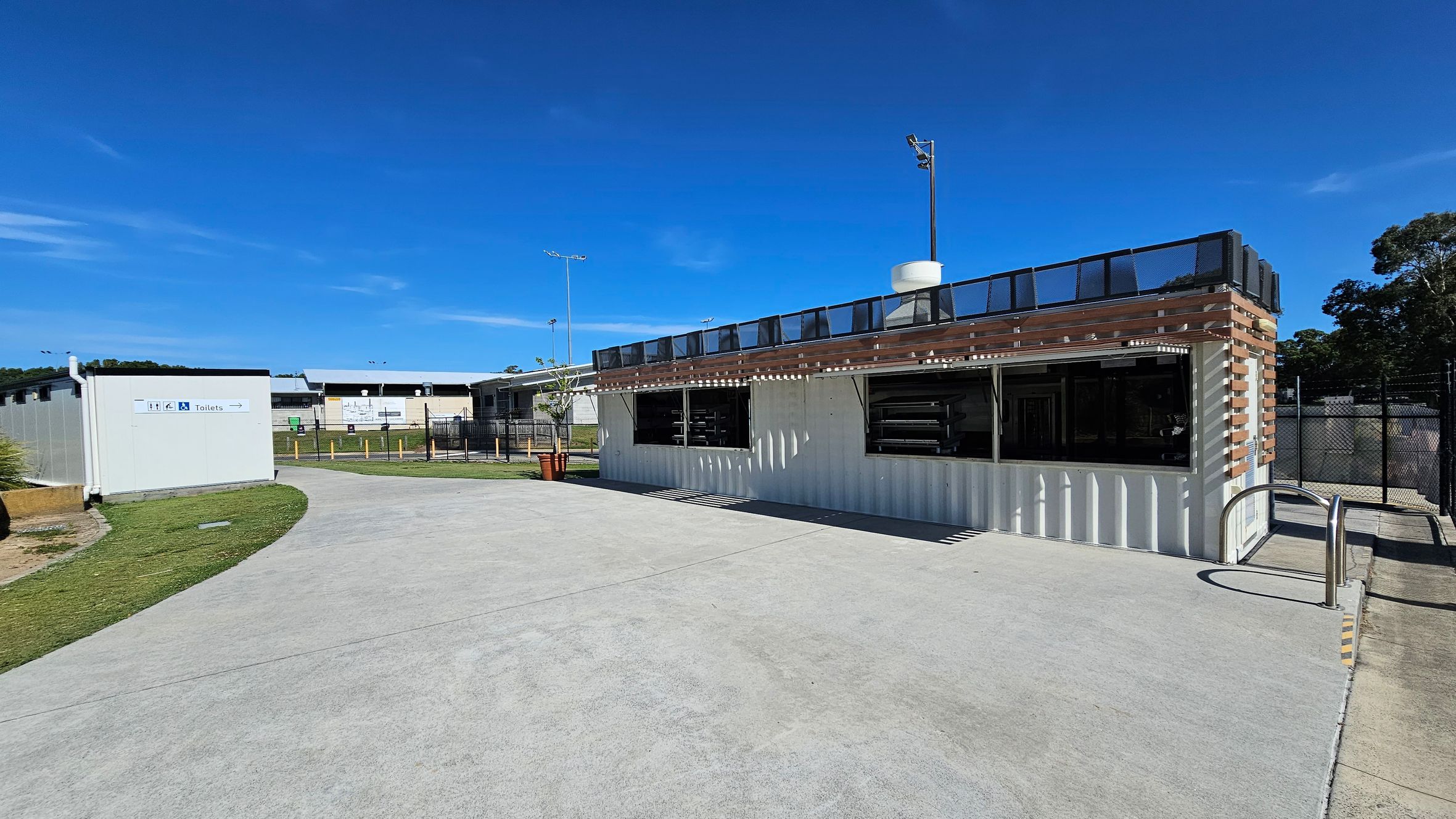 A modern building with a white exterior and wooden accents is on the right, featuring large open windows. To the left, a smaller structure with a sign indicating toilets is visible. The ground is paved with concrete, and there is a grassy area surrounding the path. In the background, there are additional buildings and a fence, with trees visible on the far right.
