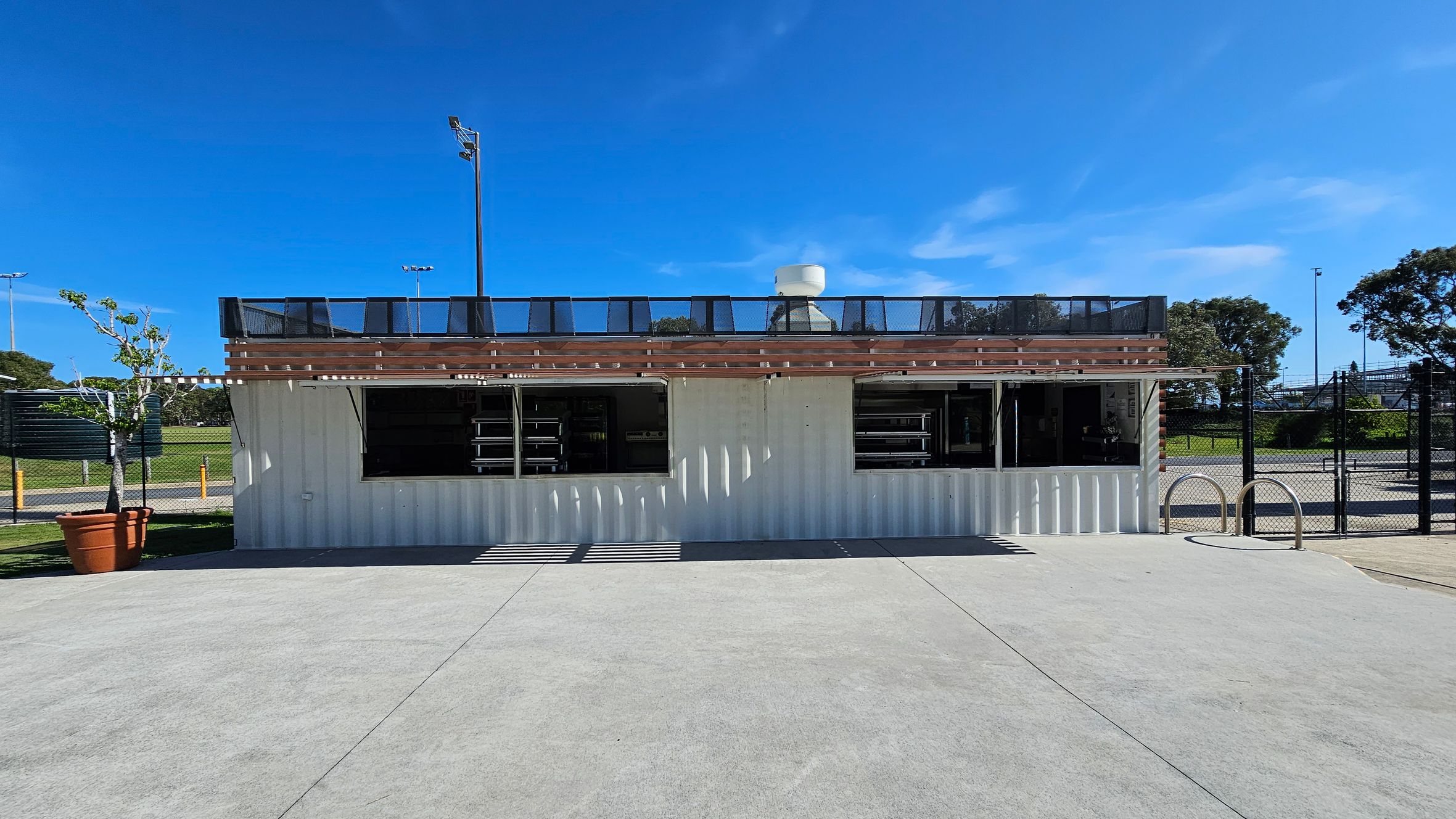 A white shipping container structure with two open windows is in the centre. It has a wooden pergola on top with a black railing. To the left, there's a potted tree. The background features a sports field with floodlights and trees. The foreground is a concrete surface, and to the right, there are metal bike racks and a chain-link fence.