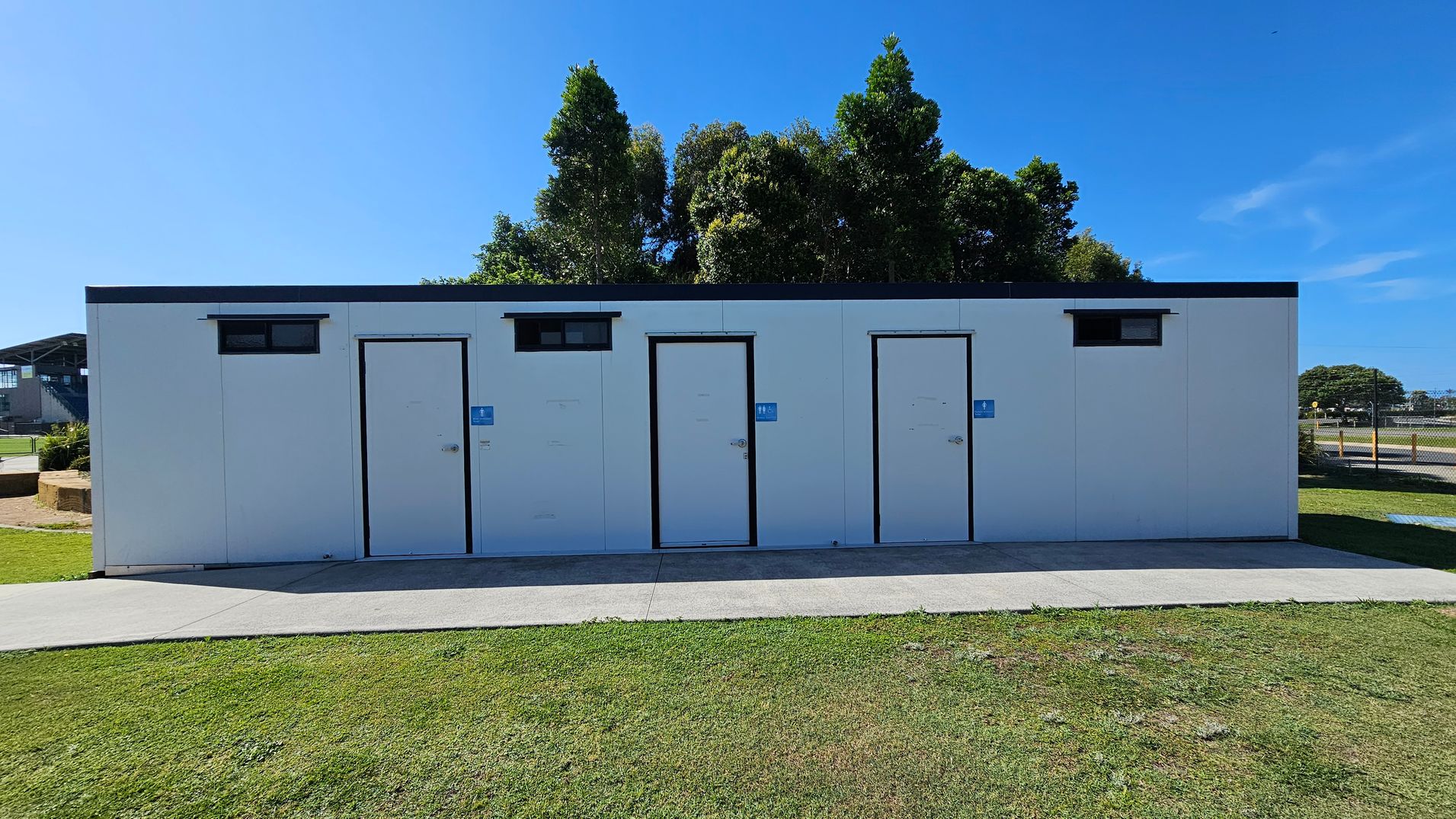 A white public toilet block with three doors, each marked with a blue sign. Small rectangular windows are above the doors. The building is set on a concrete path, surrounded by grass. In the background, there are tall trees and a glimpse of a building to the left. A fence and more greenery are visible to the right.