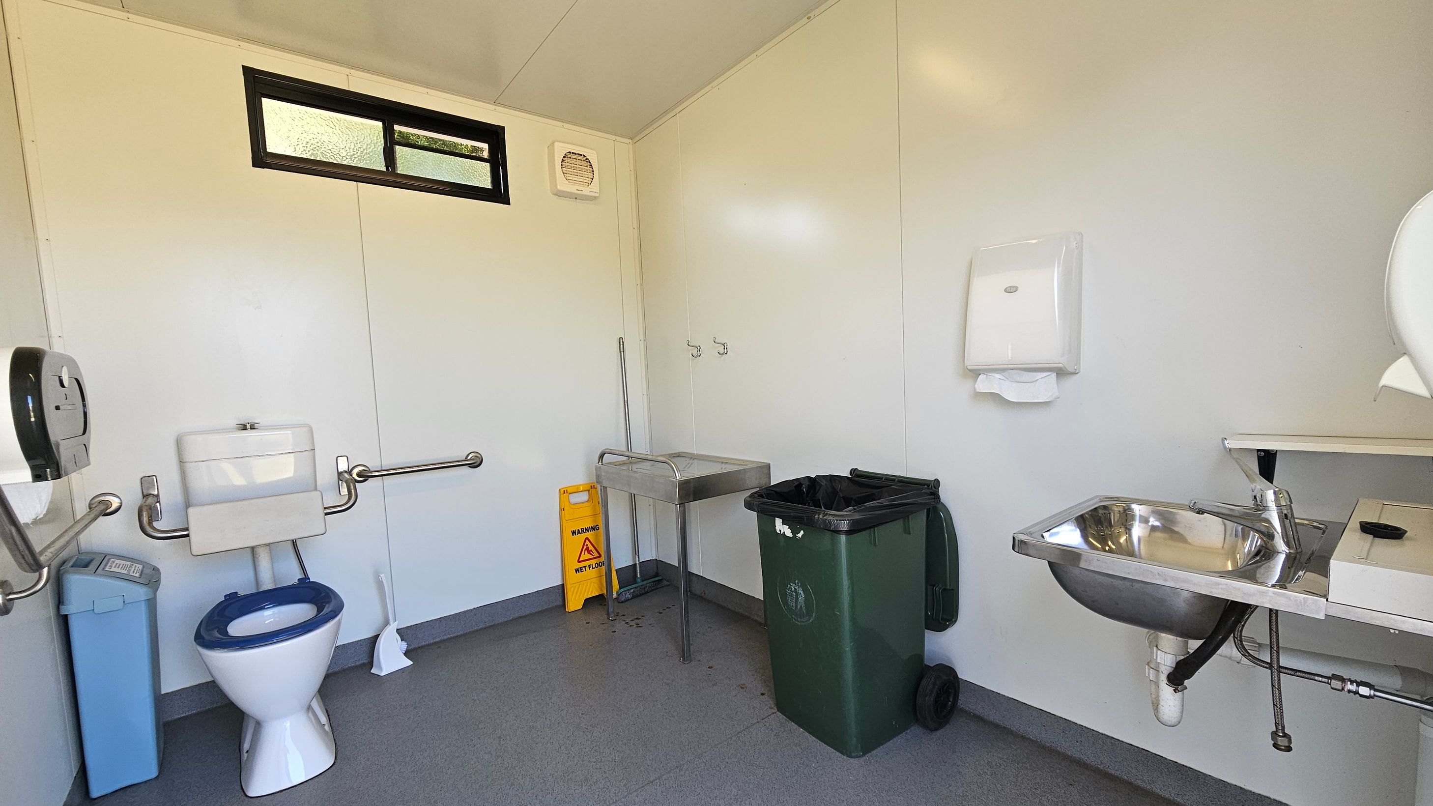 A public restroom with a white toilet featuring a blue seat on the left, equipped with grab bars. A blue sanitary bin is next to it. A small window is above. In the centre, a yellow "wet floor" sign and a mop are near a stainless steel sink. On the right, a green waste bin is beside a wall-mounted paper towel dispenser. A small metal sink with a tap is below the dispenser. The floor is grey, and the walls are light-coloured.