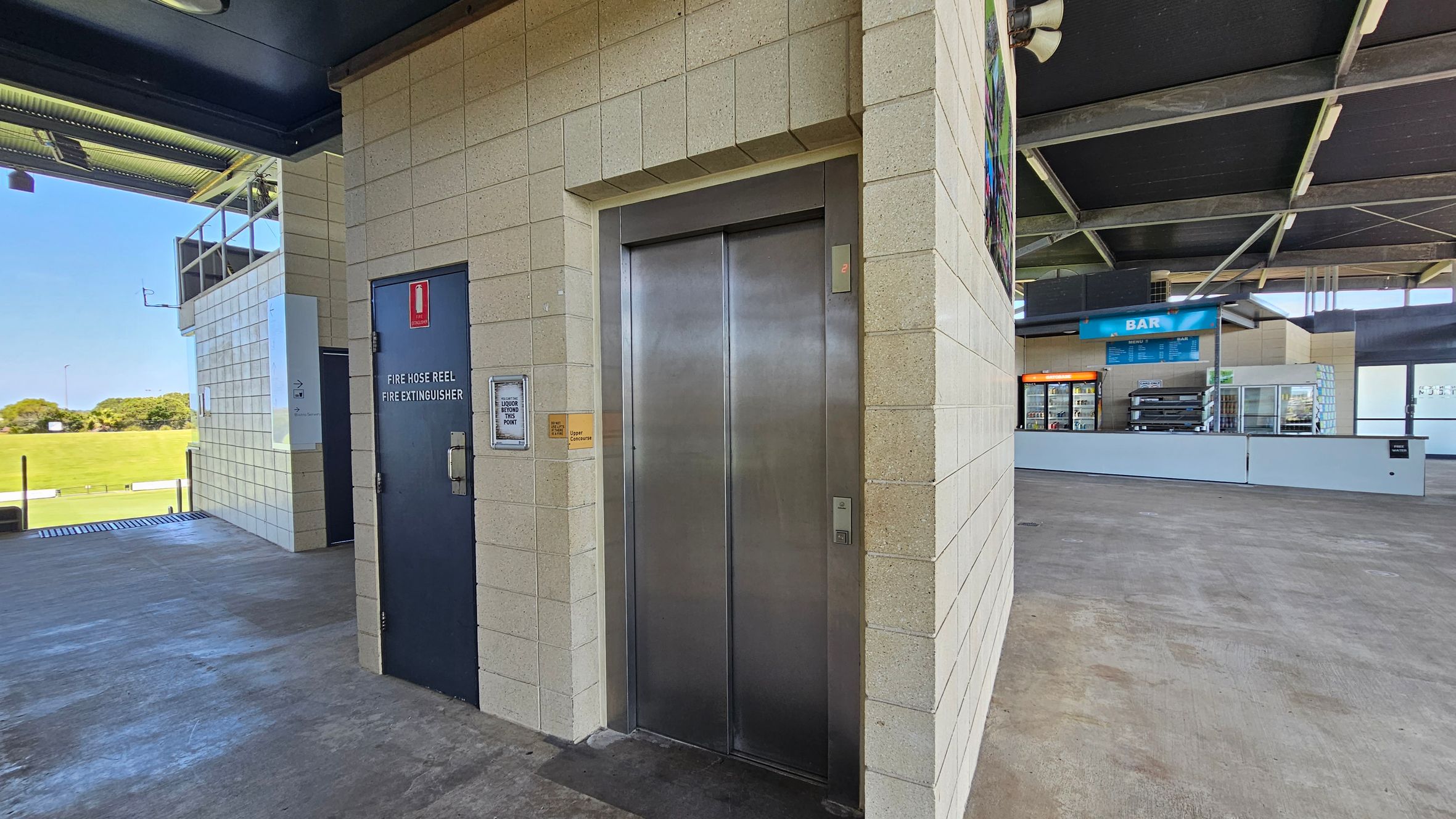 An indoor area with a concrete floor and a high ceiling. On the left, there's a dark door labelled "Fire Hose Reel Fire Extinguisher" next to a sign for a "Liquor Storage Point." In the centre, a metallic lift with closed doors is set into a light-coloured brick wall. On the right, a bar area with a blue sign, a menu, and a fridge with drinks. The background shows a grassy field visible through an open section of the building.
