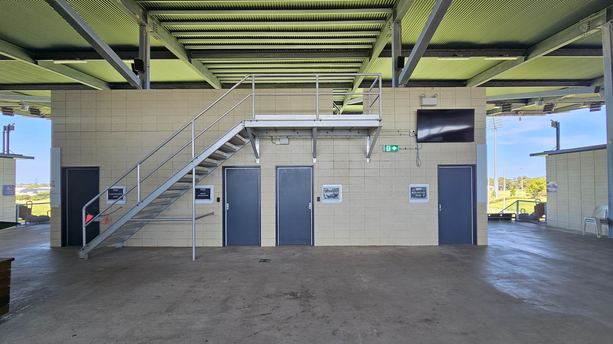 A concrete-floored area with a metal staircase leading to an upper platform. Three blue doors are set into a beige brick wall beneath the platform. A small black screen is mounted on the wall to the right. Signs and framed pictures are displayed next to the doors. The ceiling is made of metal beams and panels. Open spaces on either side reveal a grassy outdoor area with sports fields and equipment.
