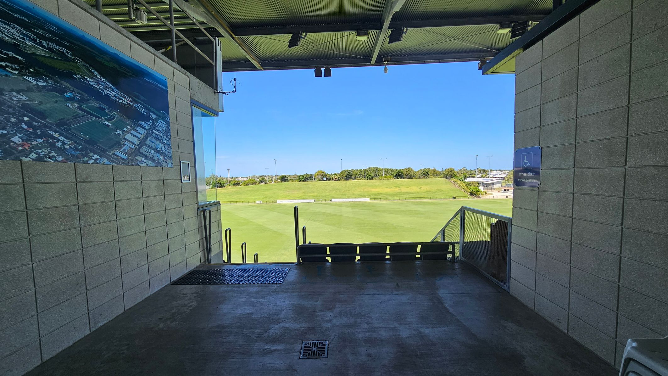 A viewing area with a concrete floor and a metal ceiling. On the left, a large aerial photograph is mounted on a brick wall. On the right, a sign indicates a wheelchair viewing area. In the middle, there are several seats facing a green sports field. Beyond the field, there are trees and a few buildings in the distance. The area is open to the outside, providing a clear view of the field.