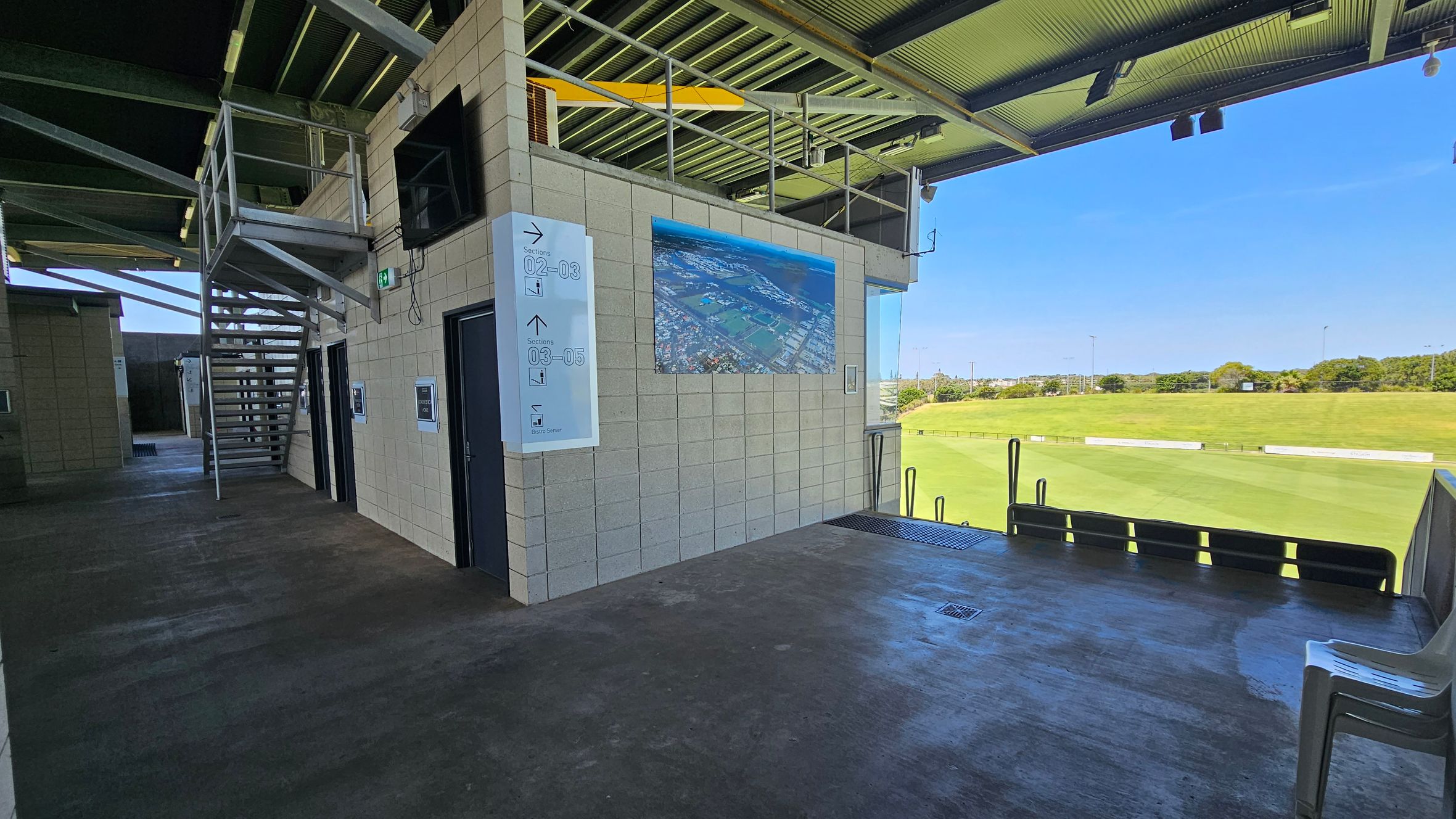 A covered outdoor area with a concrete floor and metal ceiling. On the left, there is a staircase leading up, with doors and signs on the wall. A large aerial photograph is displayed on the wall. On the right, there is a view of a green field with a few chairs placed near the edge. The area is open to the outside, allowing natural light to illuminate the space.