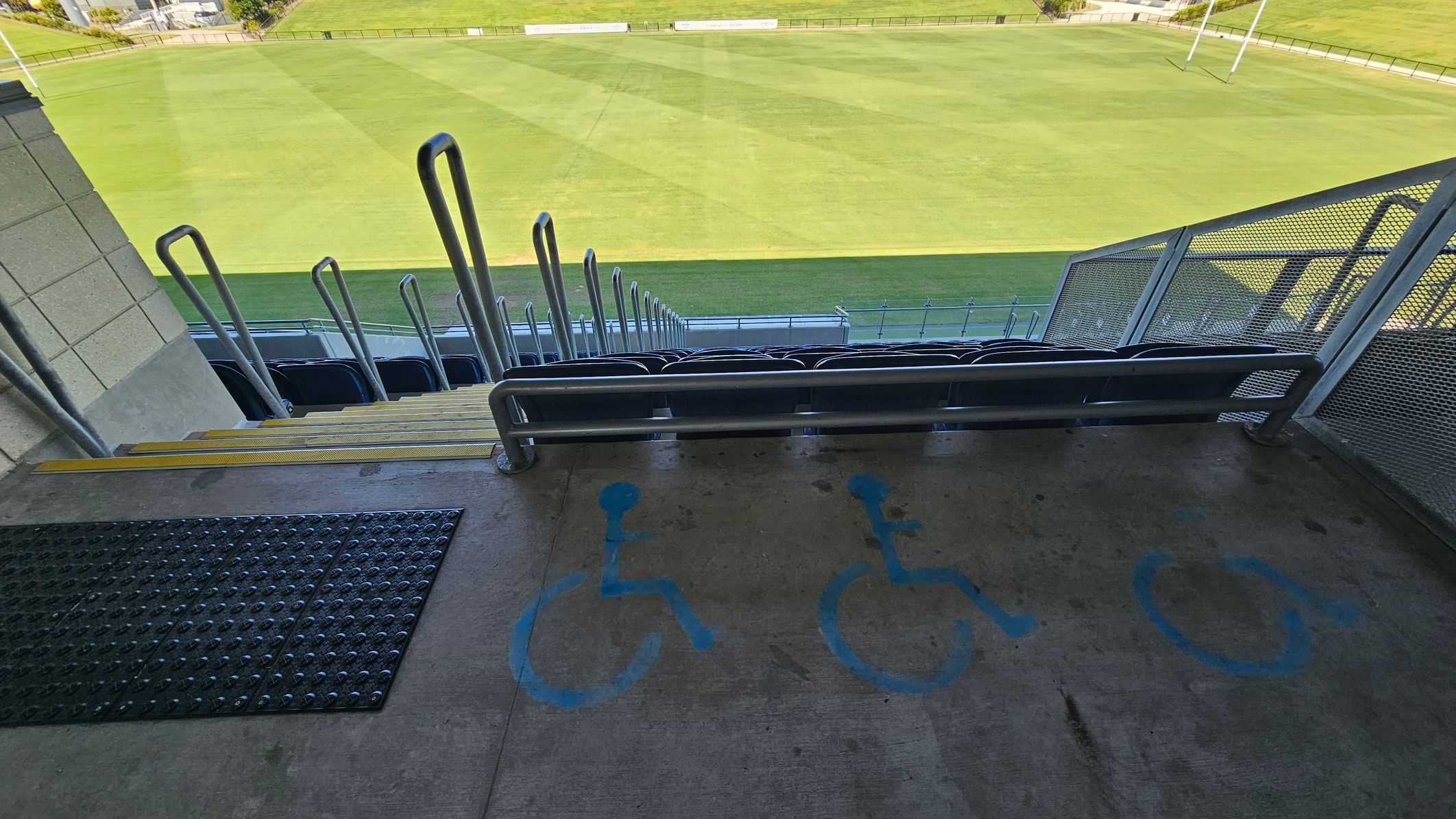 Concrete seating area with blue wheelchair symbols painted on the floor. Metal railings and steps lead down to rows of seats. Beyond the seating, a large, well-maintained grass field is visible. A mesh barrier is on the right side, and a textured mat is on the left.