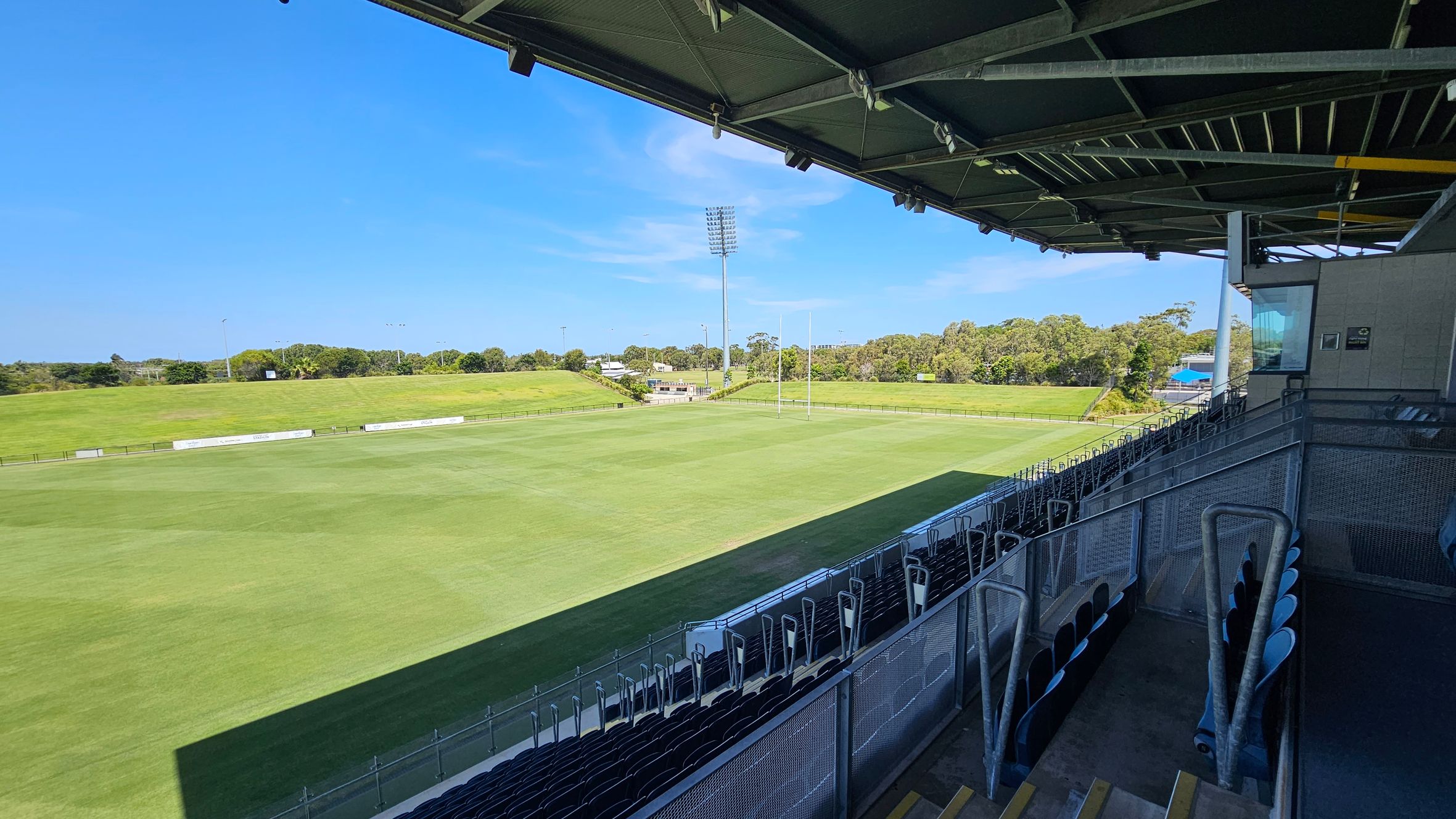 A sports stadium with a large, well-maintained grass field in the centre. On the right, there are rows of empty seats under a metal roof structure. The roof has visible beams and lighting fixtures. In the background, there are trees and a tall floodlight. The field is surrounded by a low fence, and there are advertising boards along the perimeter. The sky is clear and blue.