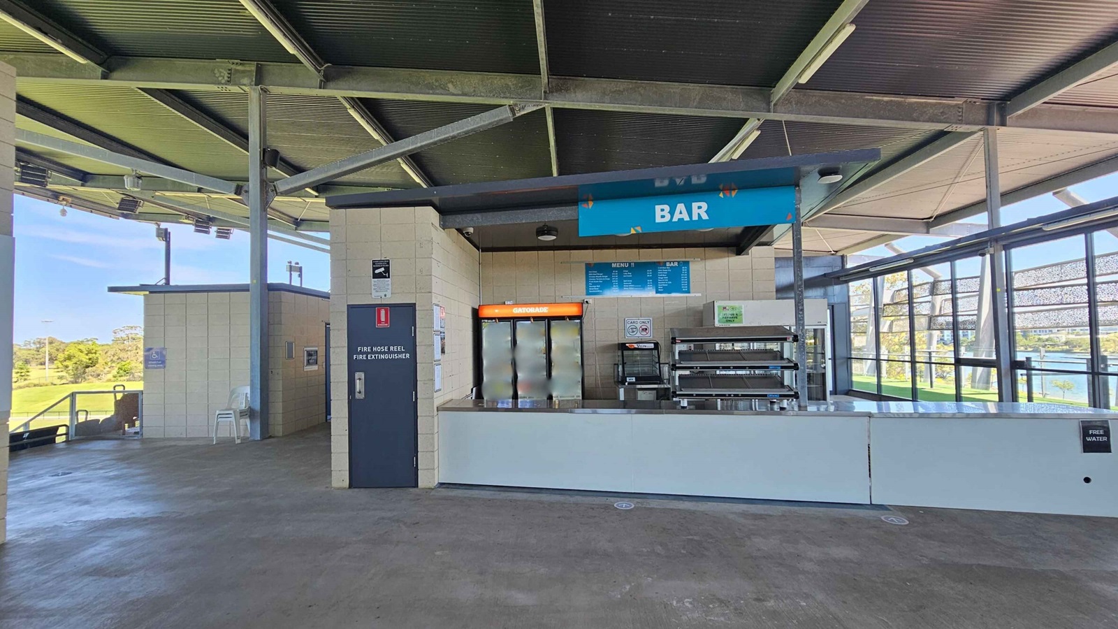 A bar area with a counter in the centre, featuring a sign above it. Behind the counter, there's a fridge stocked with drinks and a menu board. To the left, a door marked "Fire Hose Reel Fire Extinguisher" is visible, along with stacked chairs. The right side shows large windows with a view of greenery and water outside. The ceiling is made of corrugated metal, and the floor is concrete.
