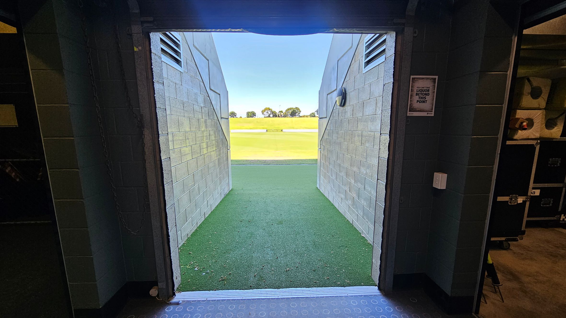 A view from inside a dimly lit corridor leading to an outdoor area. The corridor has grey brick walls on both sides, with a sign on the right wall. The floor is covered with green artificial turf extending outside. Beyond the corridor, a bright, open field is visible with a few trees in the distance. The ceiling inside is dark, and the transition from the indoor to outdoor space is marked by a metal threshold.