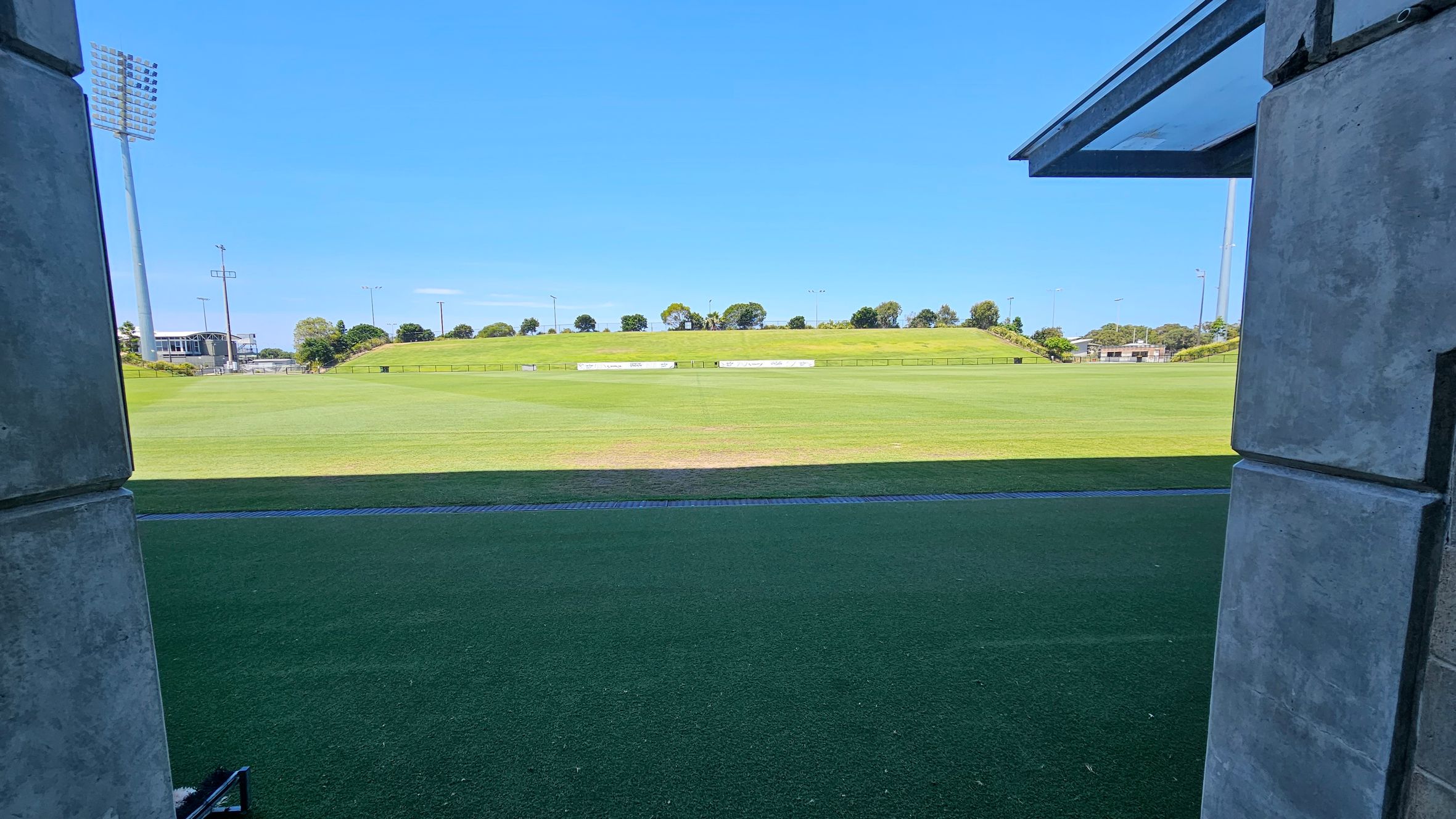 A view of a sports field from between two concrete pillars. The foreground features a strip of artificial turf leading to a large grassy field. In the background, there are trees and a small hill. Tall floodlights are visible on the left and right sides. A building is partially visible on the left, and a clear blue sky is above.