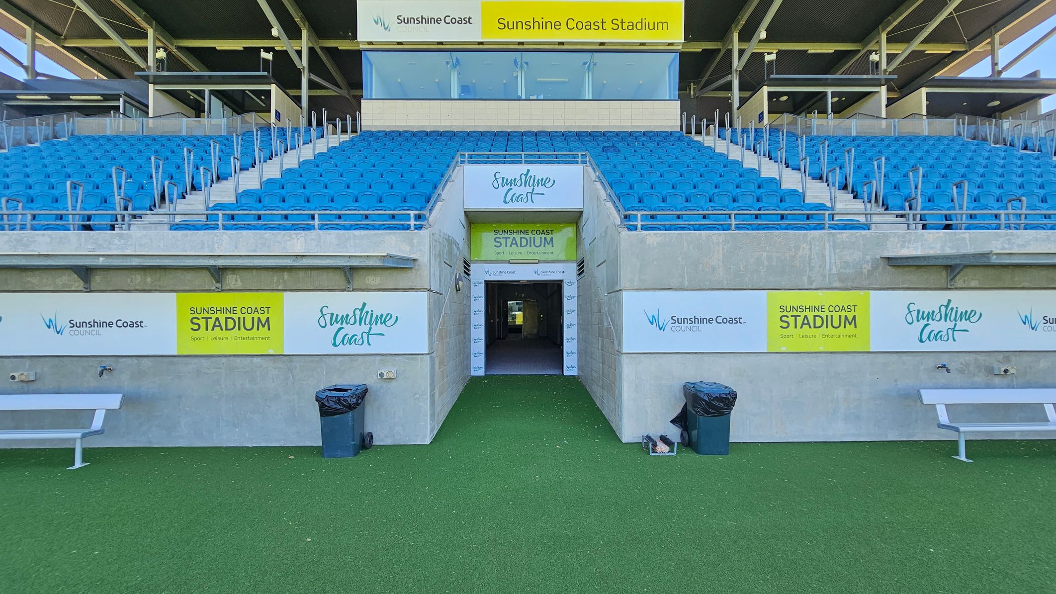 A stadium entrance with a tunnel leading through the centre, flanked by concrete walls. Above the tunnel, a sign reads "Sunshine Coast Stadium." Blue seating is arranged in rows on either side of the tunnel, with metal railings dividing the sections. The foreground features artificial green turf, a bench on the left, and two rubbish bins on either side of the tunnel. The ceiling is open with visible beams and lighting fixtures.