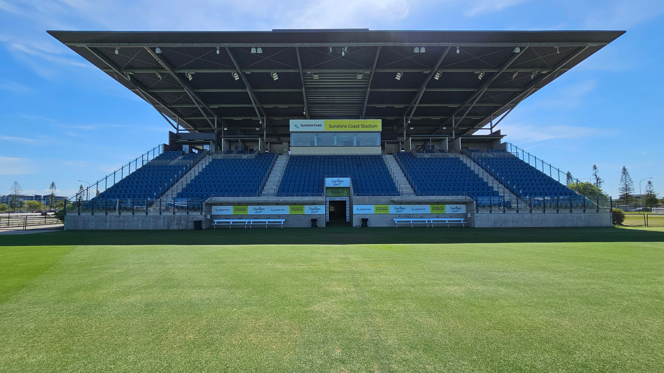 A stadium with a large, covered seating area in the centre, featuring rows of blue seats. The structure has a prominent roof supported by metal beams. Below the seating, there are entrances and signage for "Sunshine Coast Stadium." The foreground shows a well-maintained grass field. On either side of the seating, there are glass railings and staircases. In the background, there are trees and a clear sky.