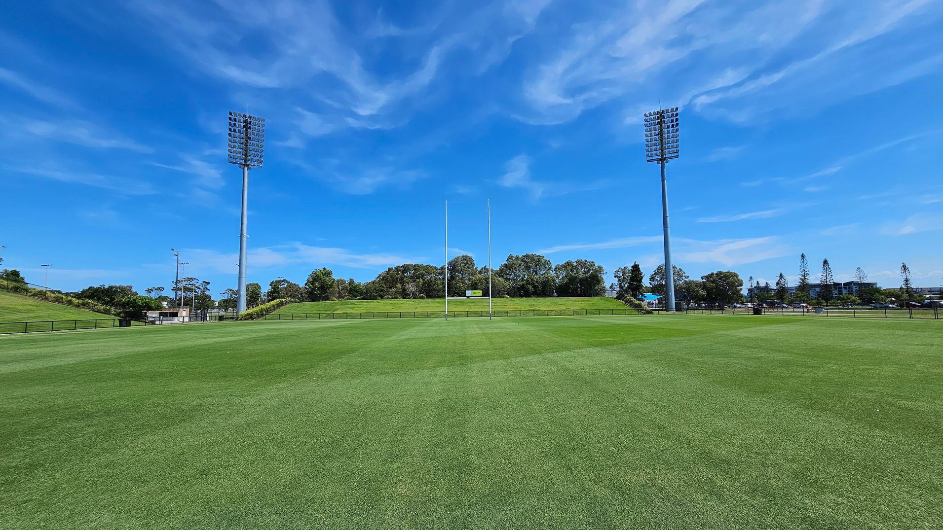 A large, well-maintained grassy sports field with two tall floodlights on either side. In the centre, there are rugby posts. The background features a low hill with trees and a small building. The field is surrounded by a fence, and there are more trees and some buildings visible in the distance.