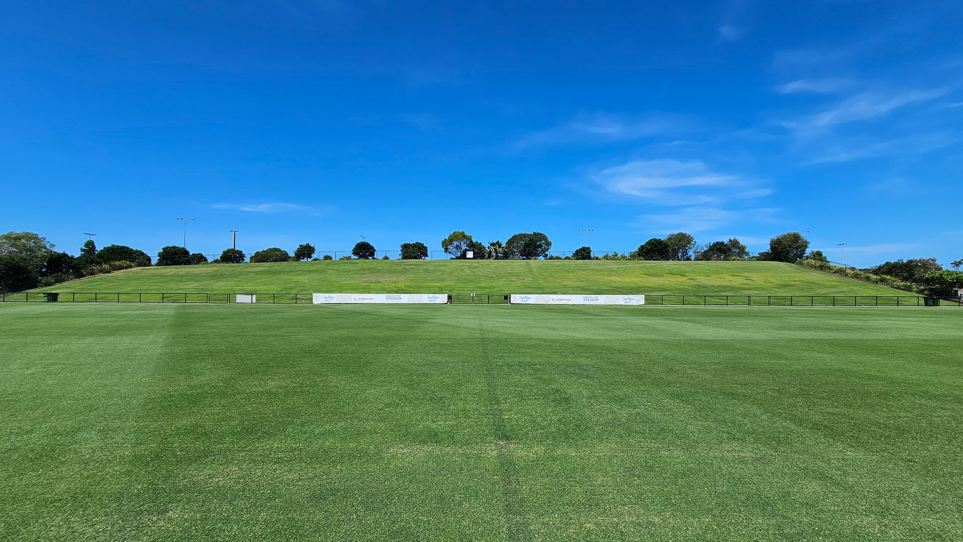 A well-maintained grassy field stretches across the foreground, leading to a gentle hill in the background. The hill is covered with more grass and lined with a row of trees at the top. There are several white advertising boards along the base of the hill. The sky is clear and blue, providing a bright backdrop to the scene.