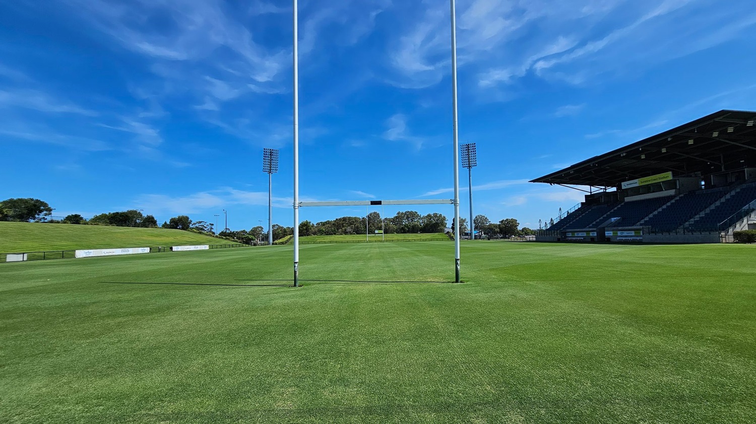 A rugby field with bright green grass. In the centre, there are rugby goalposts. On the right, a large covered grandstand with tiered seating. On the left, a grassy embankment with advertising boards. Tall floodlights are positioned on both sides of the field. Trees and shrubs are visible in the background.