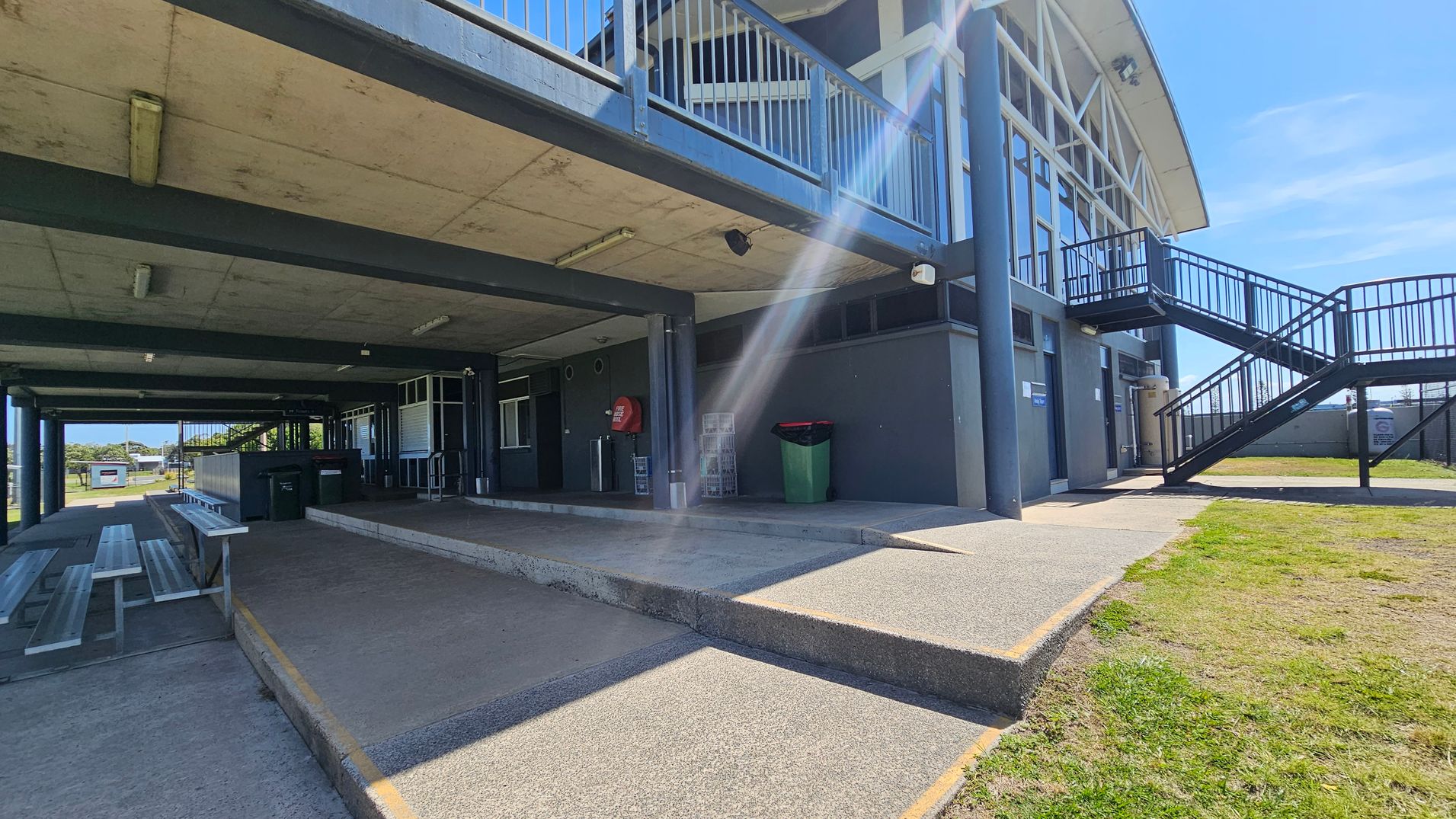 A two-storey building with a concrete overhang and metal railings. On the left, there are benches and a pathway leading to a sports field. In the centre, a ramp and steps lead to a covered area with a green bin and a red fire hose reel. On the right, an external staircase ascends to the upper level. The ground is a mix of concrete and grass, and the ceiling has fluorescent lights.