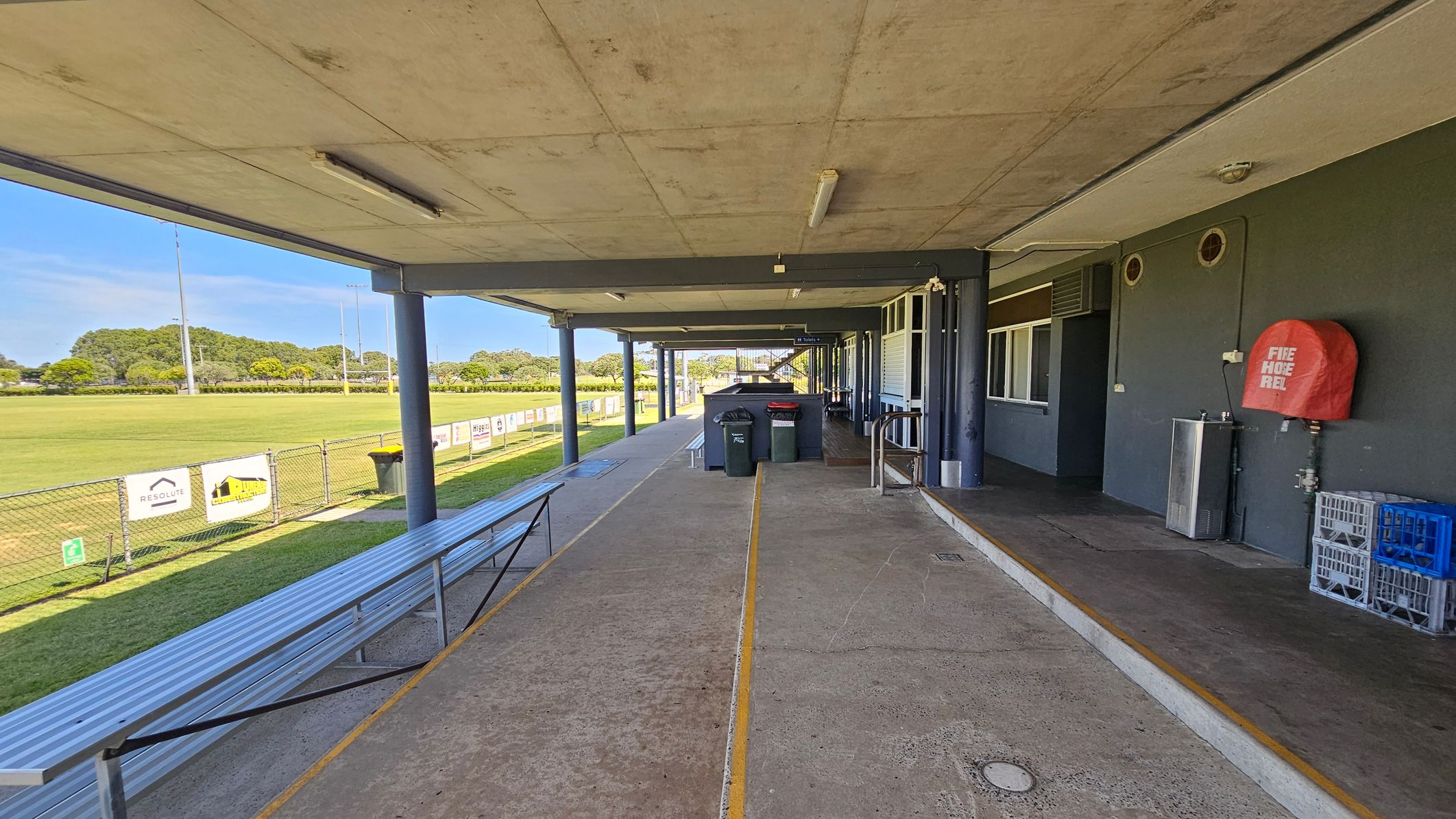 A covered walkway with a concrete floor and ceiling. On the right, there's a dark wall with a red fire hose reel, a drinking fountain, and stacked crates. In the centre, bins are placed along the path. On the left, a metal bench runs parallel to a chain-link fence, beyond which is a grassy field with advertising signs. The area is supported by blue-grey pillars.