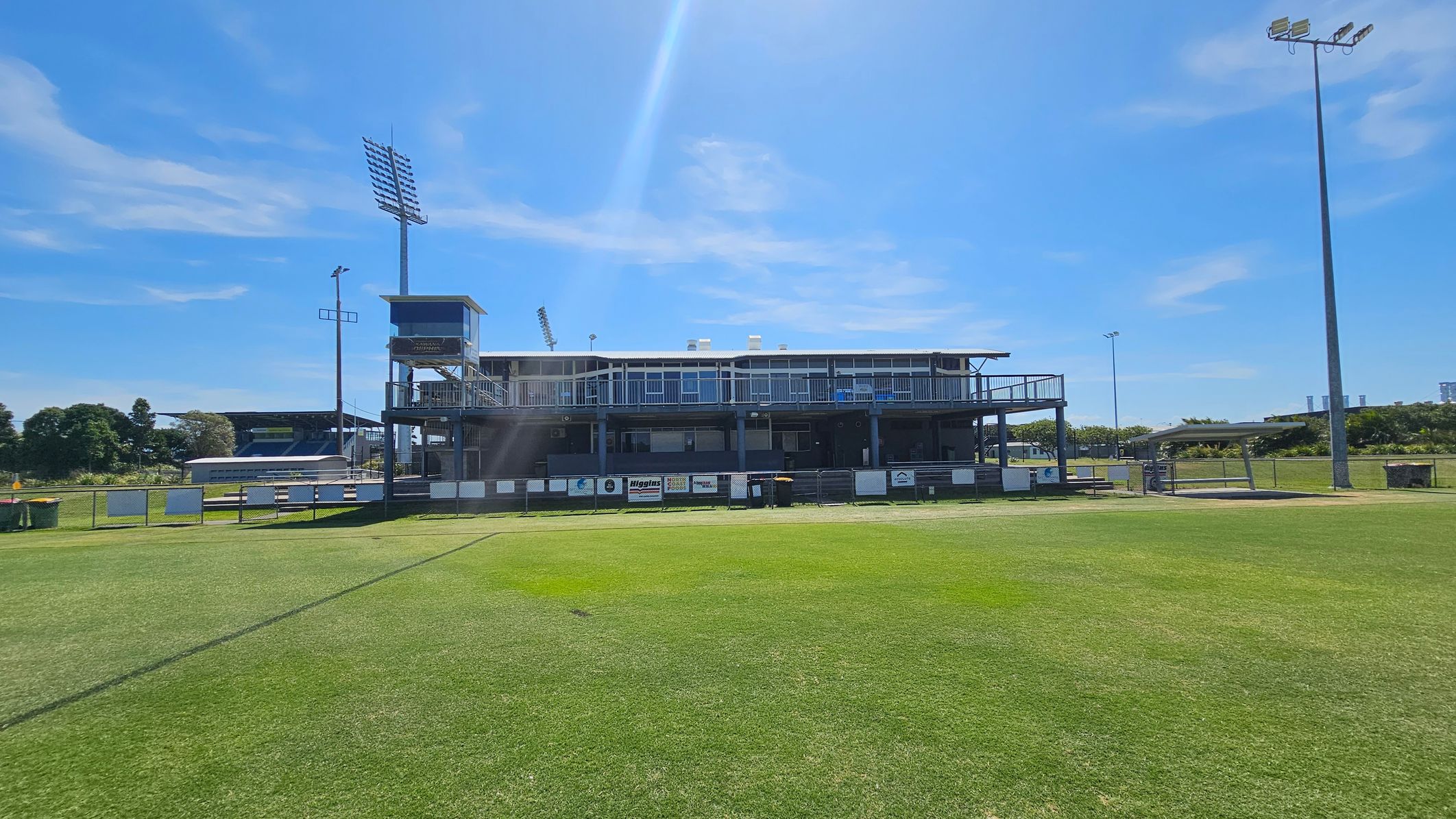 A sports field with a large, two-storey building in the centre, featuring a balcony and multiple windows. The building is surrounded by a fence with various signs and advertisements. On the left, there is a tall floodlight and a smaller structure. The grass is well-maintained, and there are a few trees and additional structures in the background. The sky is clear and blue.