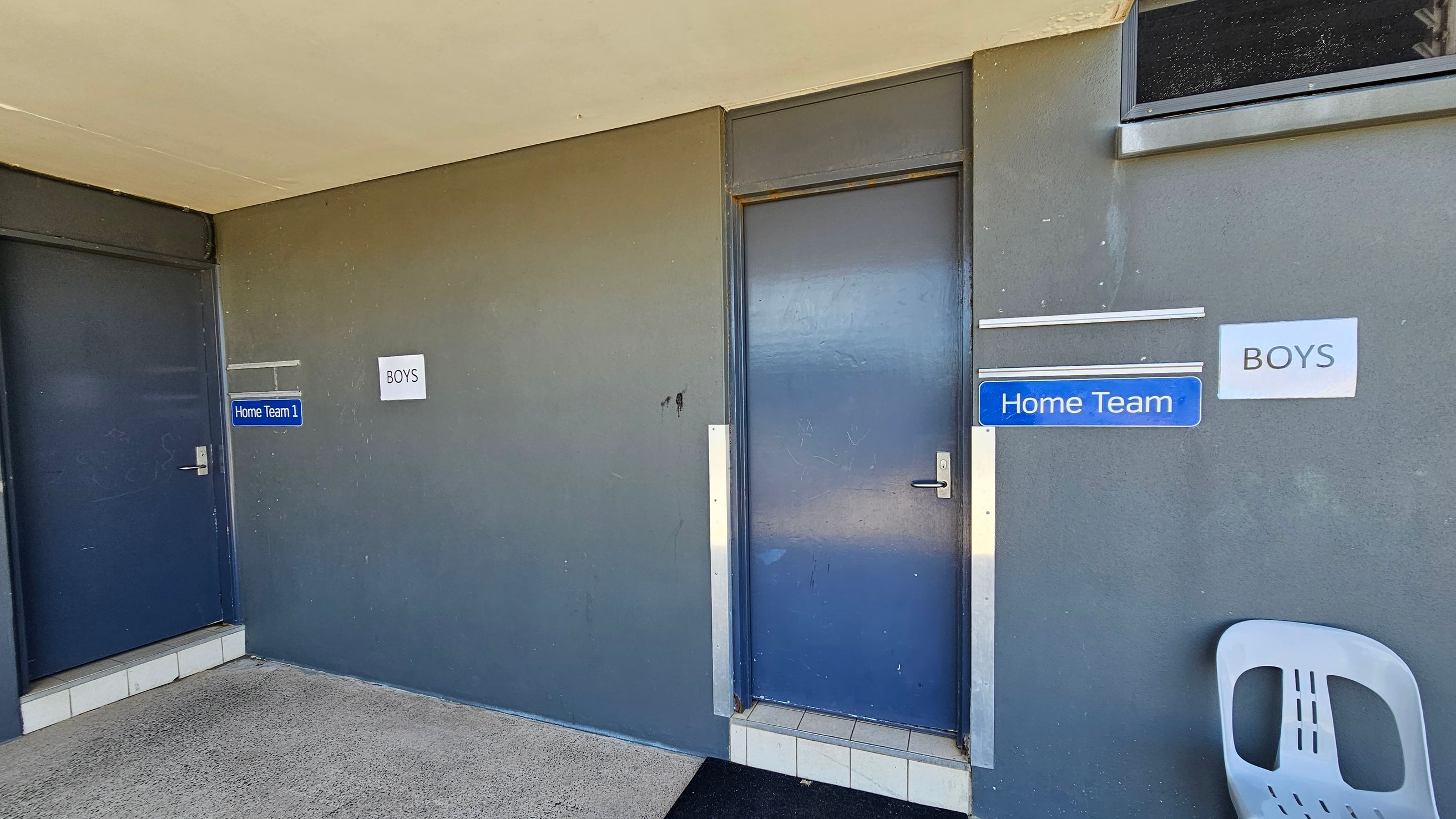 A corridor with two blue doors on a grey wall. The left door has a sign reading "Home Team 1," and the right door has signs for "Home Team" and "Boys." A grey plastic chair is positioned to the right of the second door. The floor is concrete, and the ceiling is light-coloured.