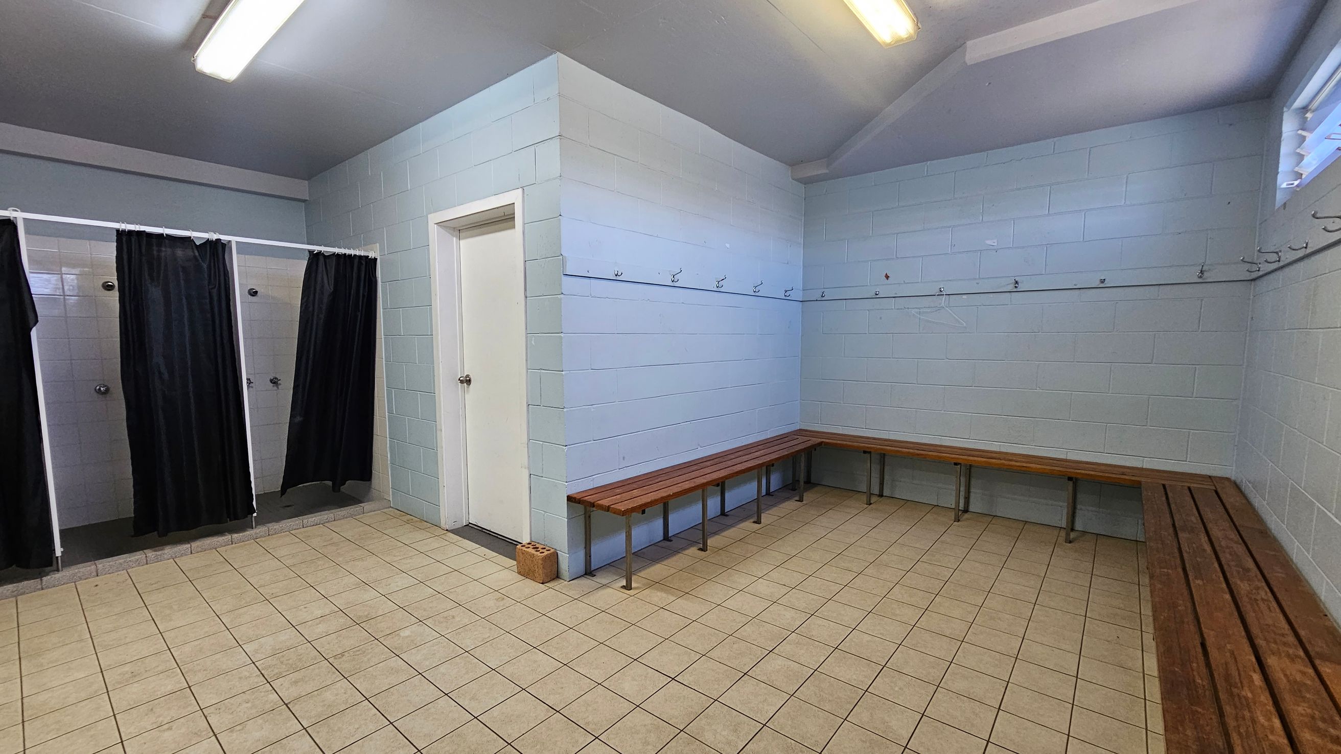 A changing room with light blue and white tiled walls. On the left, there are three shower stalls with black curtains. In the centre, a white door is visible. On the right, wooden benches line the walls, with hooks above them for hanging items. The floor is covered with beige tiles, and the ceiling has fluorescent lights. A small window is on the upper right wall.
