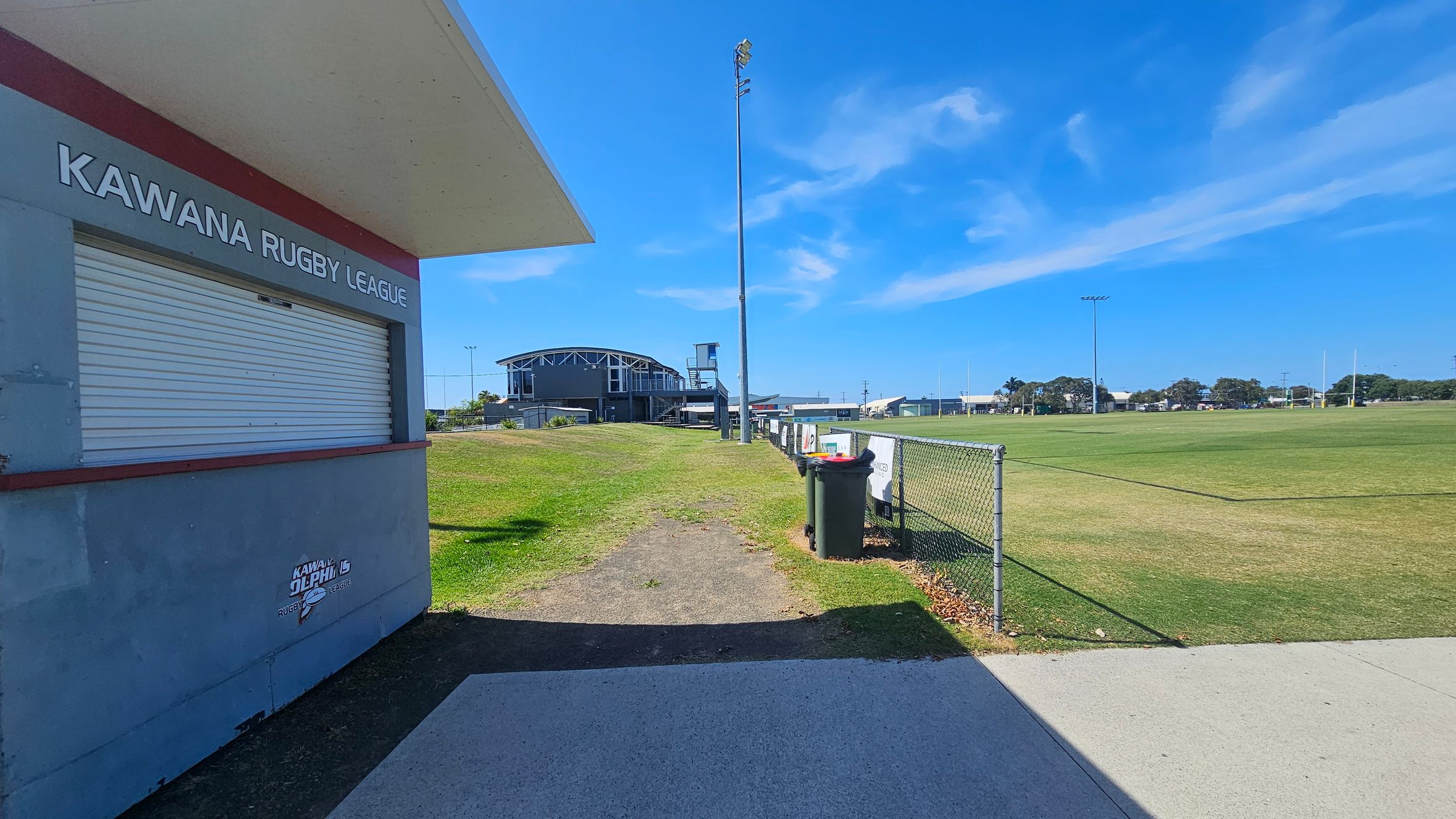 On the left, a building with a closed shutter and the words "Kawana Rugby League" is visible. In the middle, a grassy area leads to a large sports field with a tall floodlight. A fence runs alongside the field, with bins placed near it. In the background, a large building with a distinctive roof structure is seen. The ground is a mix of concrete and grass.