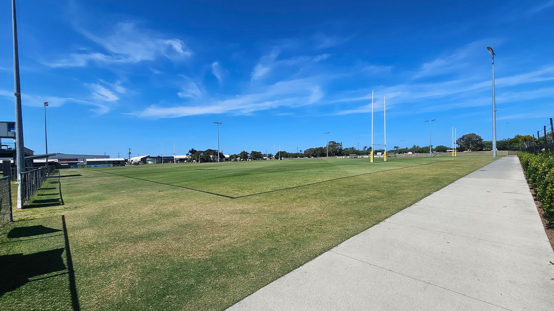 A rugby field with green grass and goalposts in the centre. On the left, there is a fence and a building in the distance. On the right, a concrete path runs alongside the field, bordered by a hedge. Tall floodlights are positioned around the field.