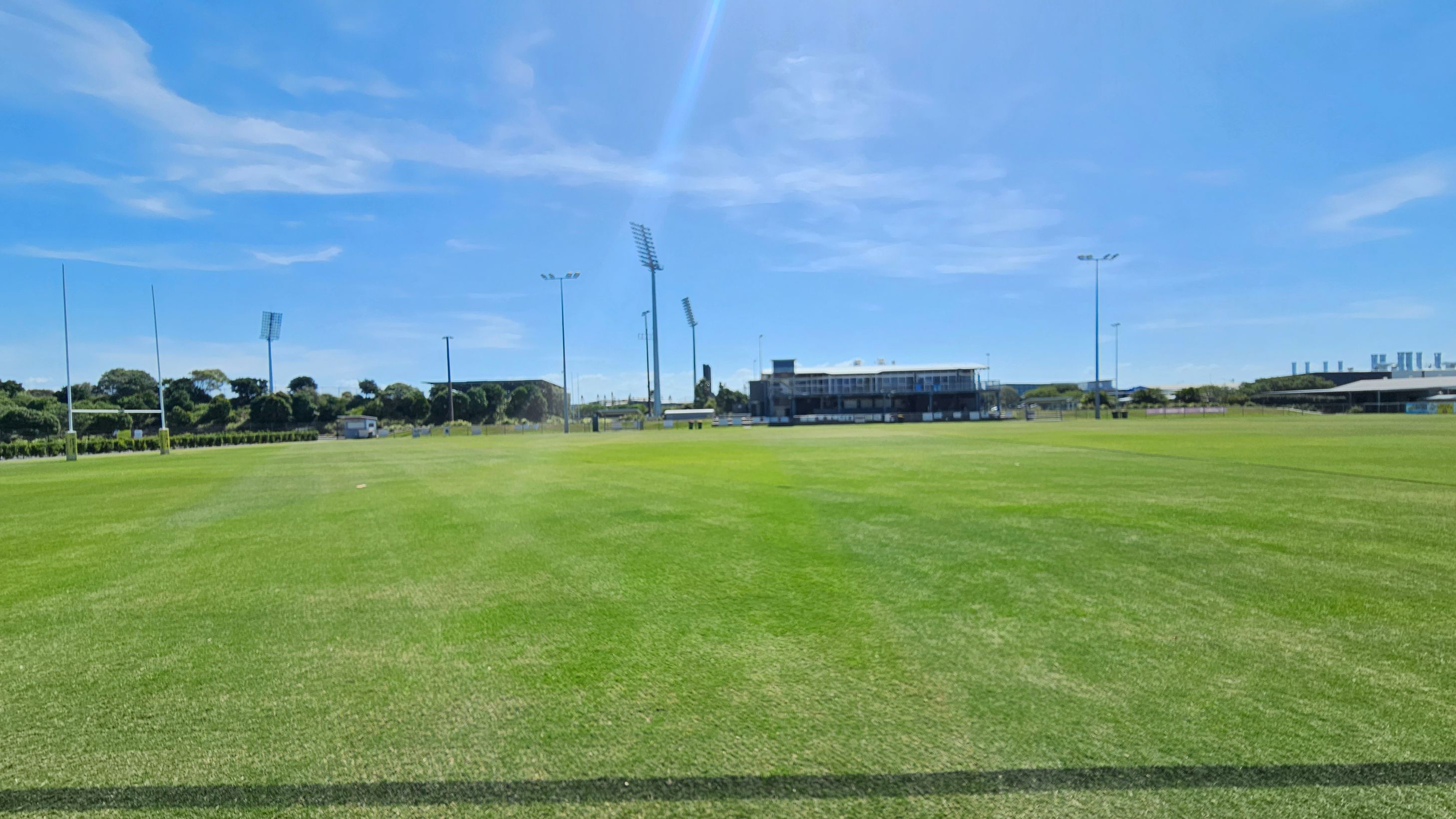 A large, well-maintained grassy sports field with rugby posts on the left. In the background, there are several tall floodlights and a building with a viewing area. Trees and some industrial structures are visible in the distance. The field is expansive, with clear lines marking the playing area.